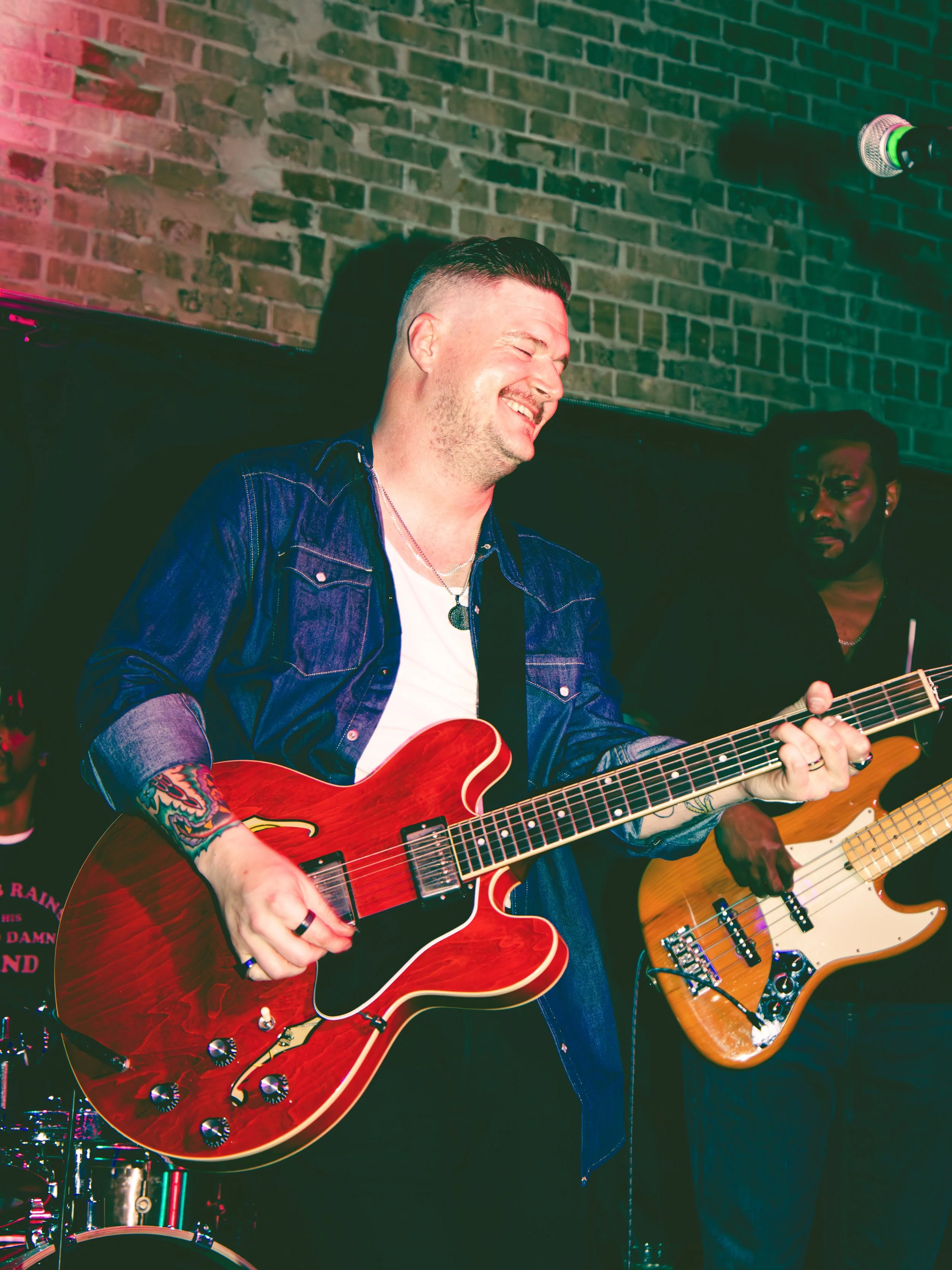 A man with a short haircut and tattoos on his left arm, wearing a denim jacket and a white T-shirt, is smiling while playing a red acoustic guitar. Another man with glasses and a beard is in the background playing a yellow electric bass guitar. They 