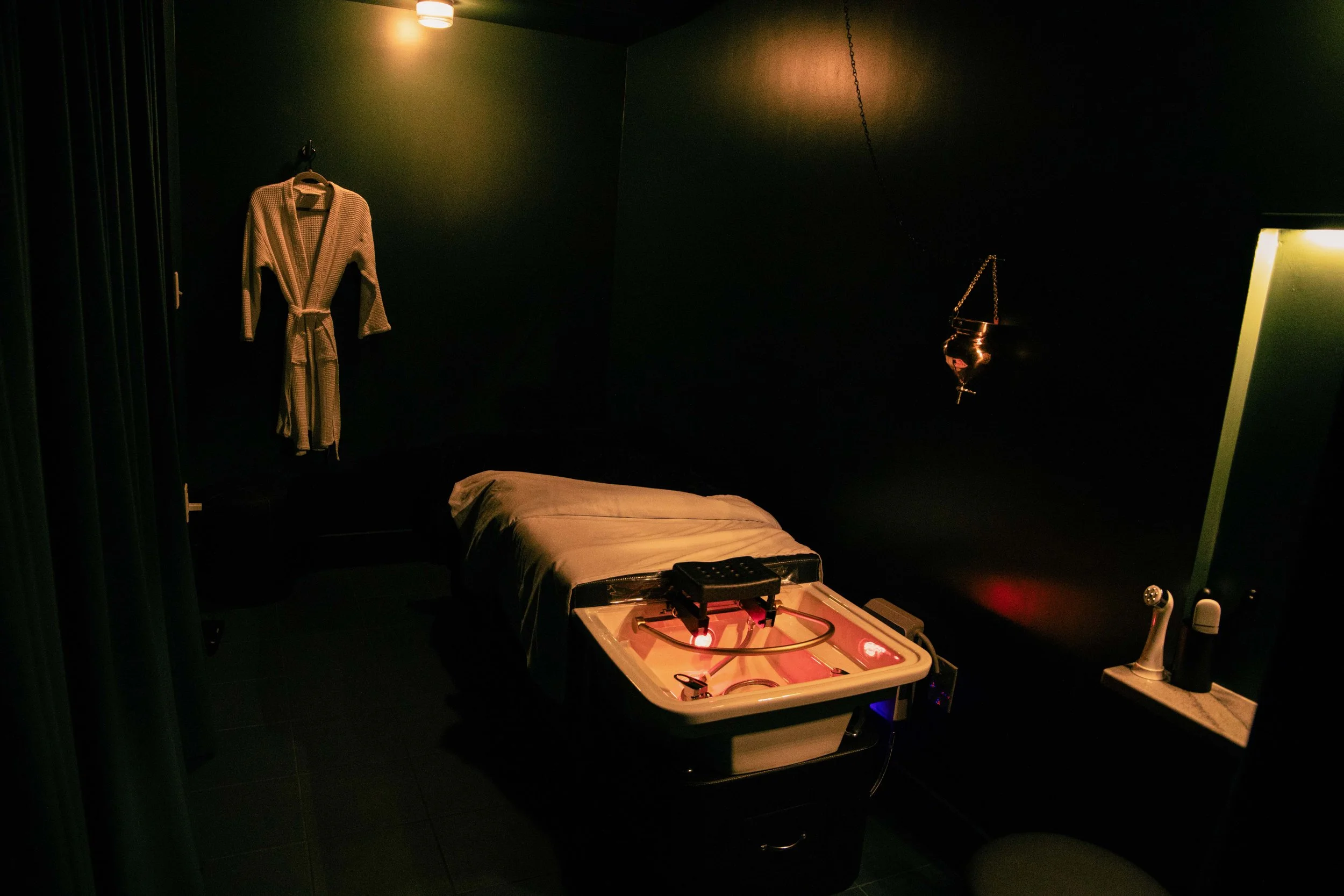 A dimly lit room with dark green walls. There is a bed with a beige blanket, a small table with medical or scientific equipment, a hanging light, and a robe hanging on the wall. A hanging metal object and a partial mirror or screen are also visible.