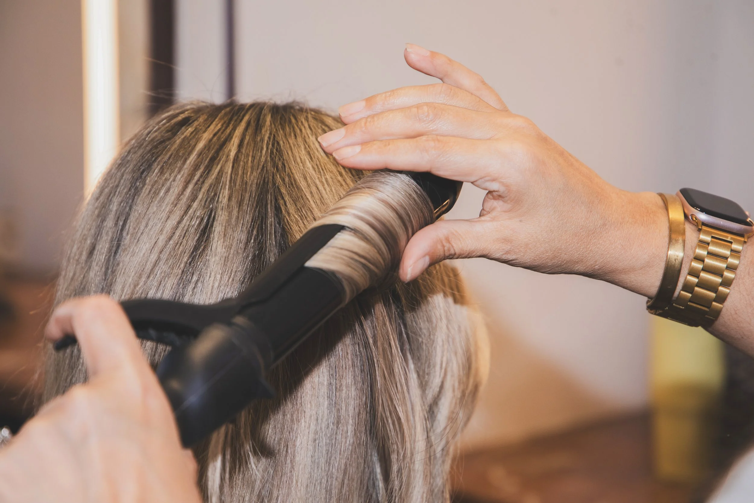 A person is using a flat iron to straighten a section of a woman's long, blonde hair in a salon.