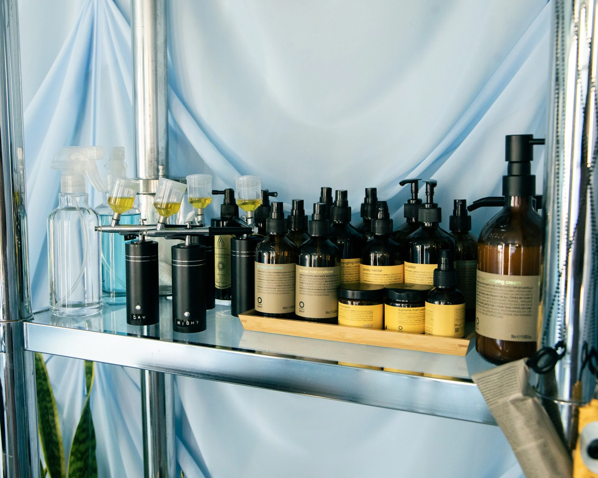 A metal shelf containing various bottles and containers with black and transparent spray nozzles and gold labels, along with some small transparent bottles with yellow liquid, set against a blue curtain backdrop.