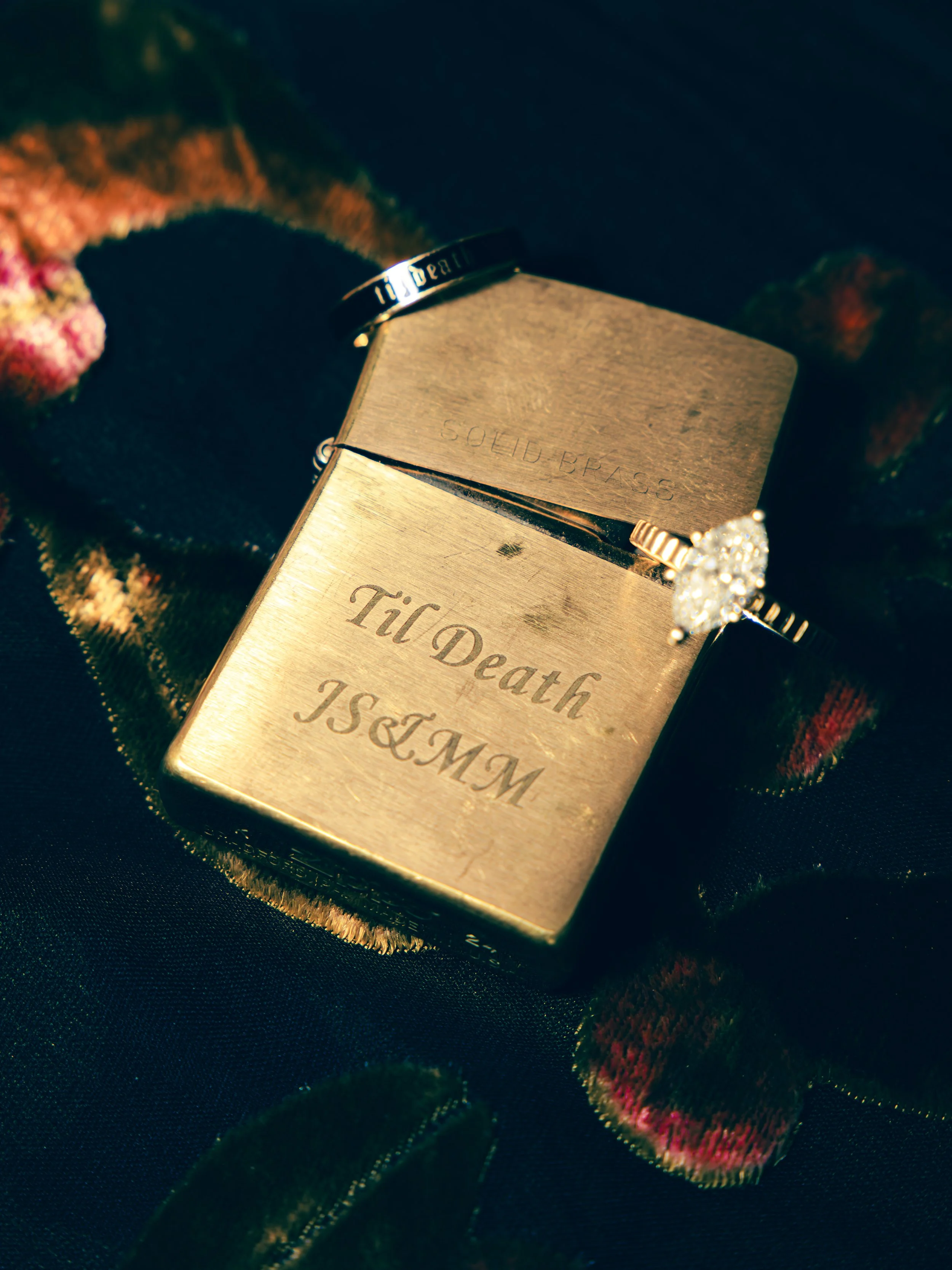 A gold-colored box with the inscription 'Till Death' and a diamond ring resting on top of it, surrounded by dark fabric with red and green velvet flowers.