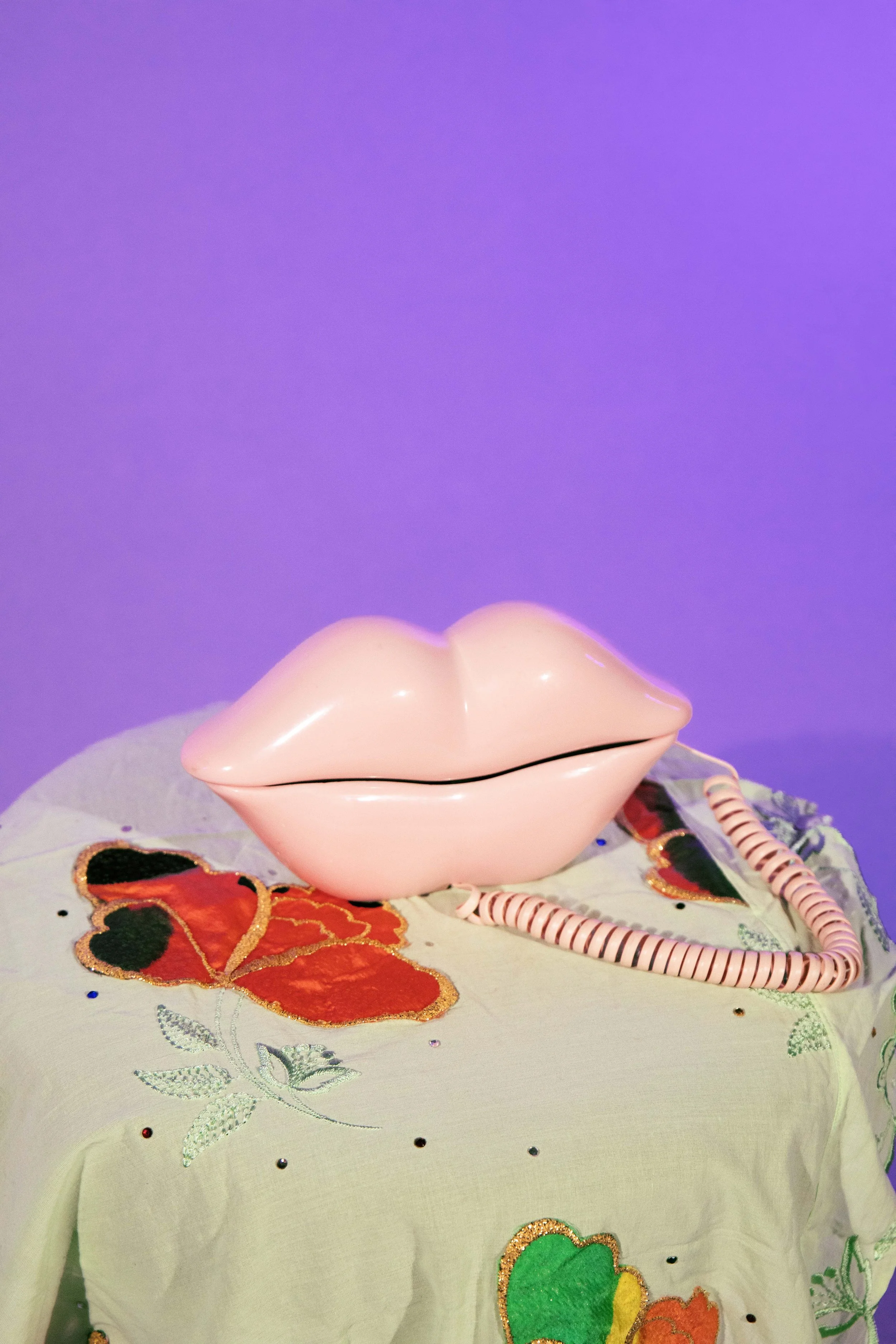 A pink lip-shaped ceramic container with a lid, placed on a decorative embroidered fabric with floral patterns and colorful accents, against a purple background.