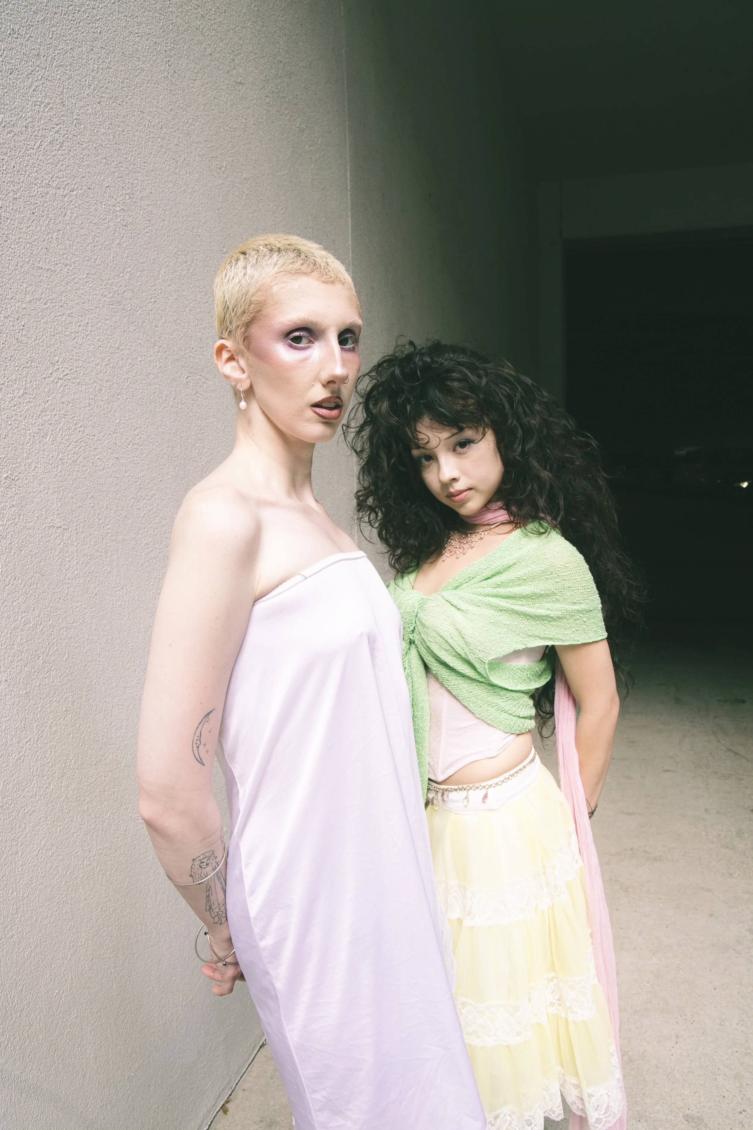Local Dallas, Texas, DFW Non-traditional Fashion Editorial Creative Portraits Cover Art Photographer Photoshoot. Two women standing close together against a plain wall. The woman on the left has short blonde hair, light skin, and is wearing a white s