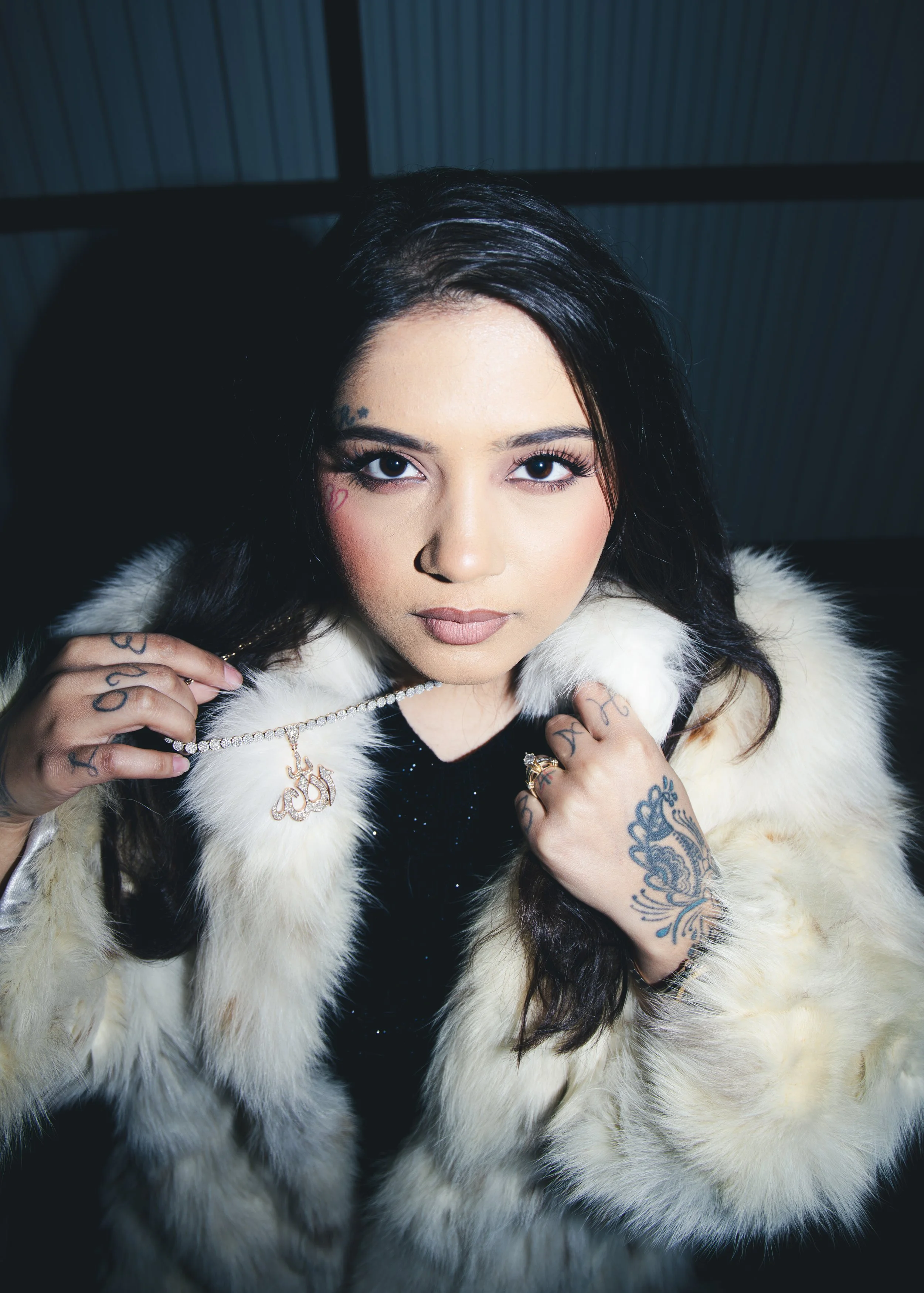 Local Dallas, Texas, DFW Non-traditional Fashion Editorial Creative Portraits Cover Art Photographer Photoshoot. Close-up of a woman with dark hair, tattoos, and makeup, wearing a white fur coat, holding a diamond necklace with a pendant, and looking