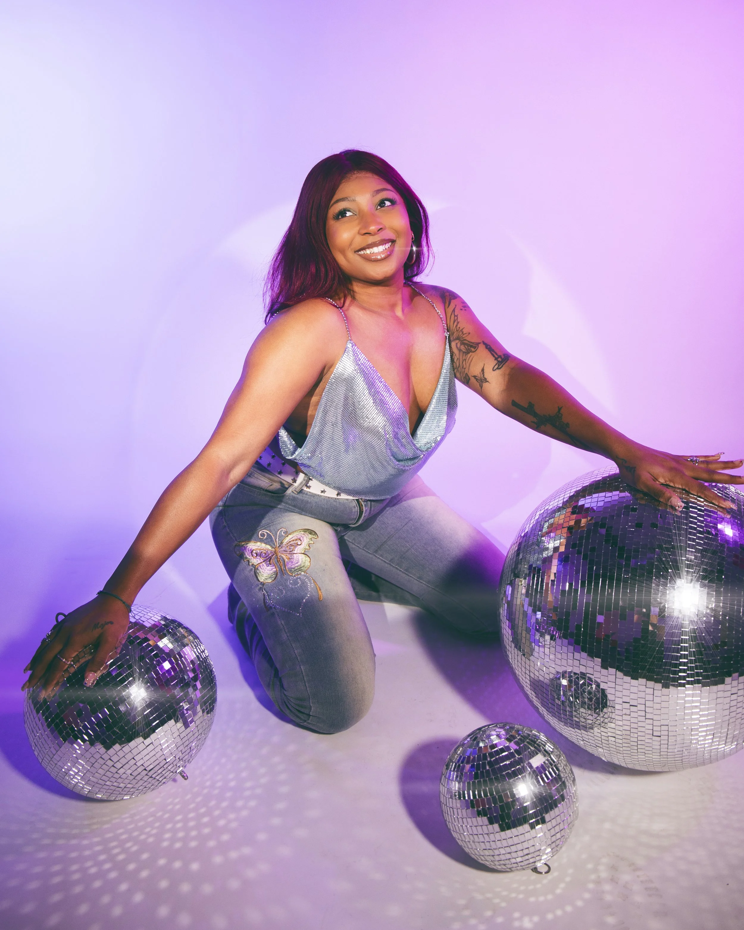 Local Dallas, Texas, DFW Non-traditional Fashion Editorial Creative Portraits Cover Art Photographer Photoshoot. Musician Seihara .A woman kneeling in front of four disco balls on a white surface with purple lighting, smiling and looking upward.