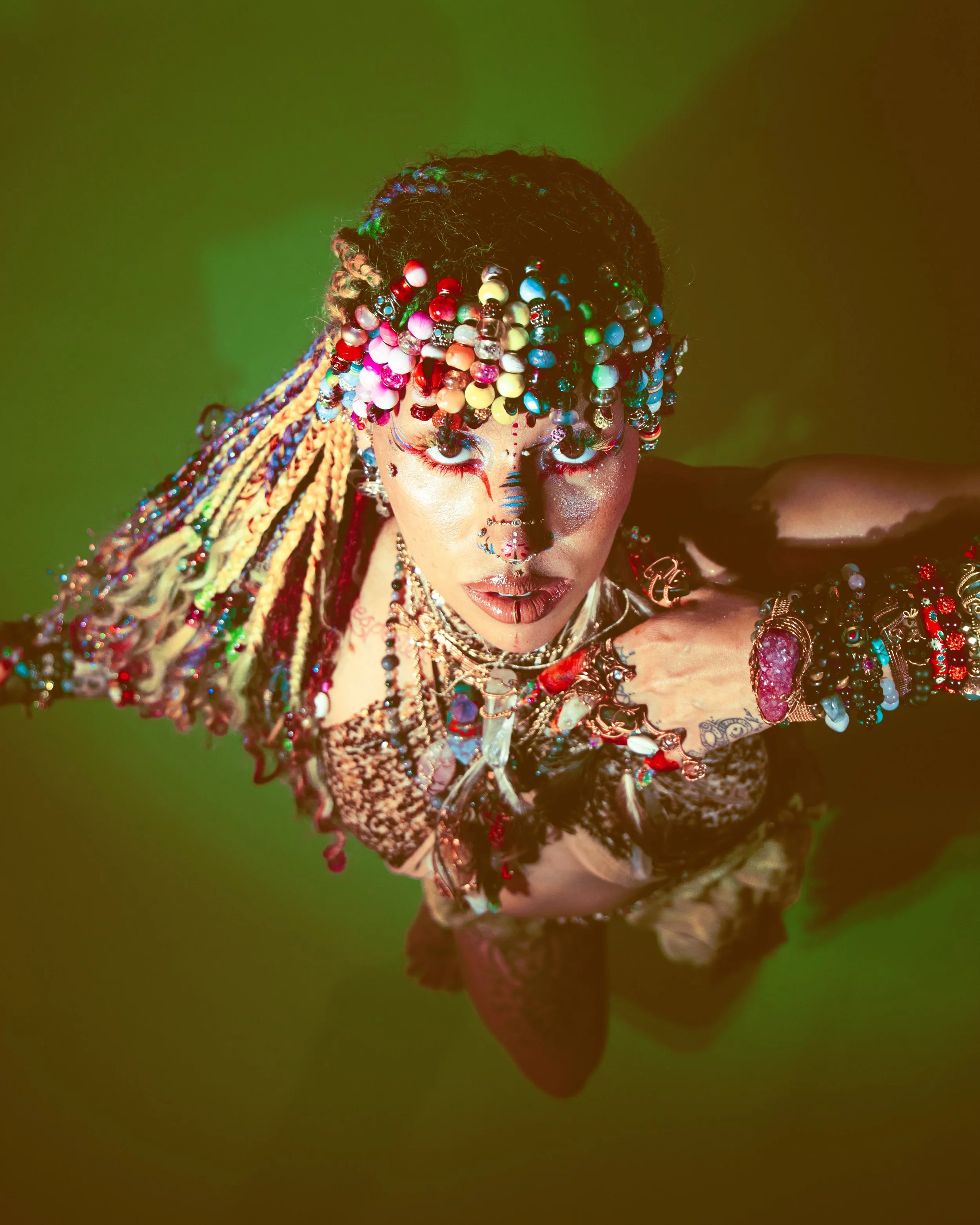 Local Dallas, Texas, DFW Non-traditional Fashion Editorial Creative Portraits Cover Art Photographer Photoshoot. A person with colorful beads and jewelry in a dramatic pose with face painted, wearing elaborate makeup and accessories, against a green 
