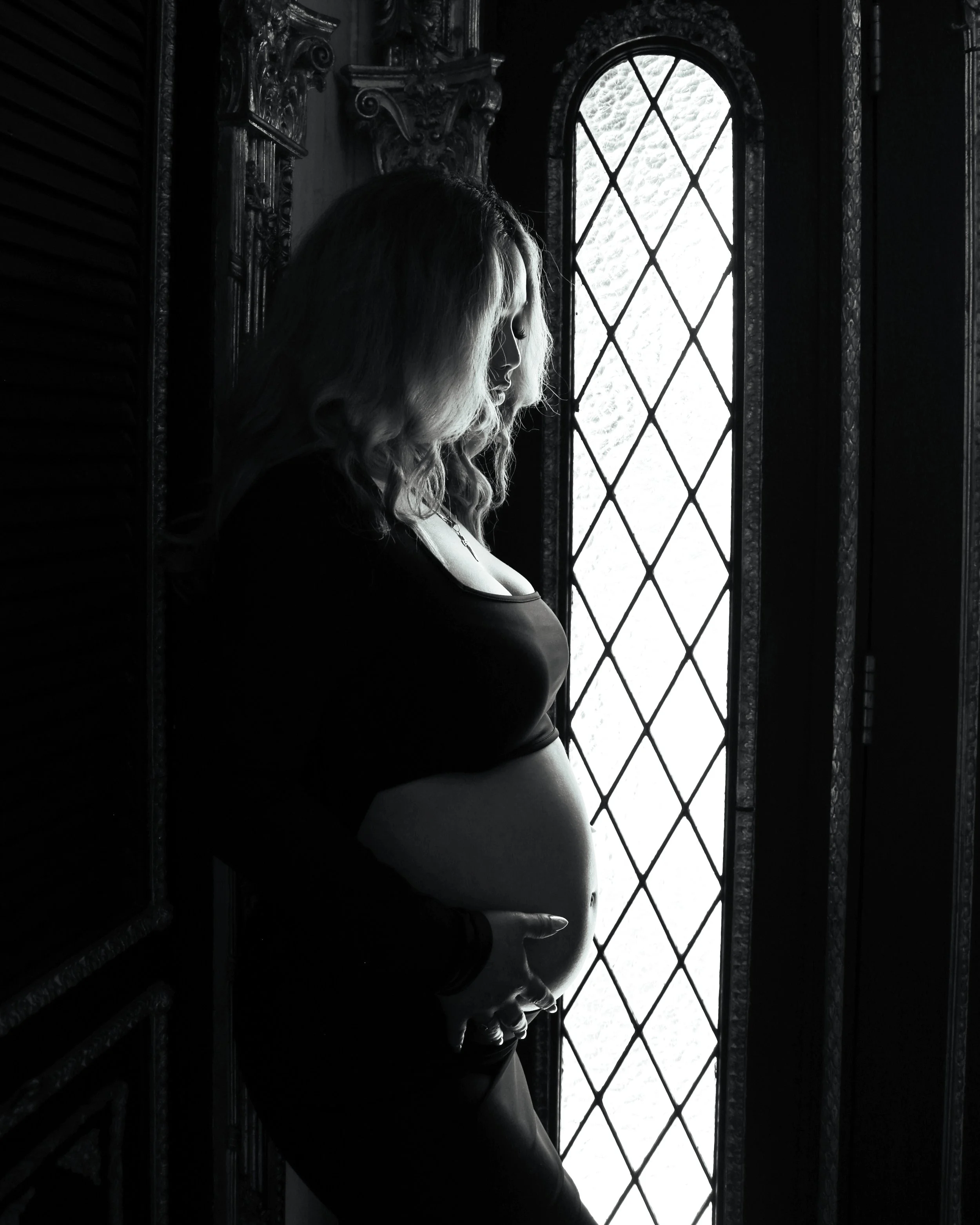 A black and white photo of a woman with long, curly hair looking down, standing in front of a decorative window with diamond-shaped panes, inside a dark, ornate room.