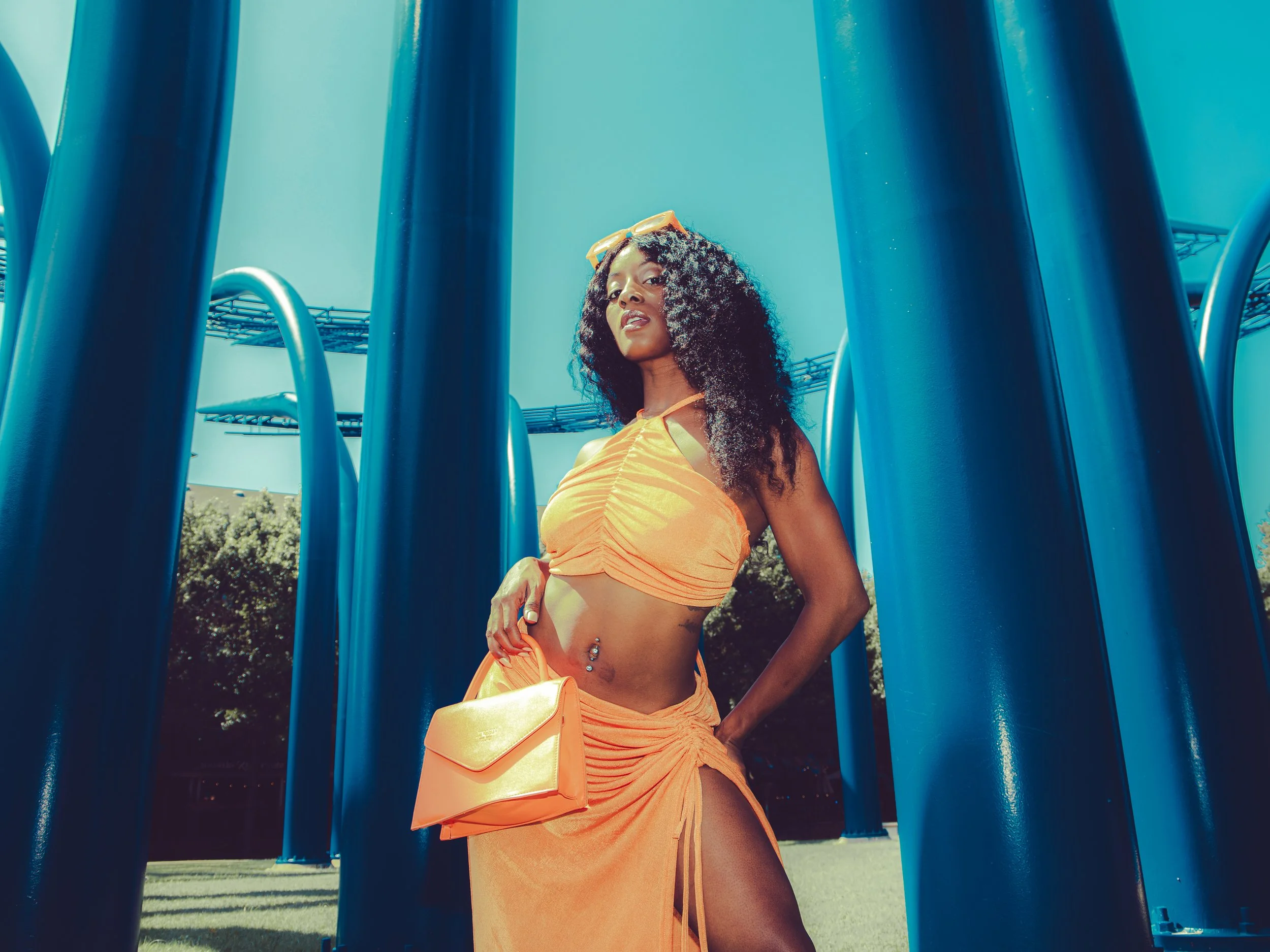 Local Dallas, Texas, DFW Non-traditional Fashion Editorial Creative Portraits Cover Art Photographer Photoshoot. A woman wearing orange clothing and sunglasses on her head stands beside blue playground equipment. Addison Circle. 