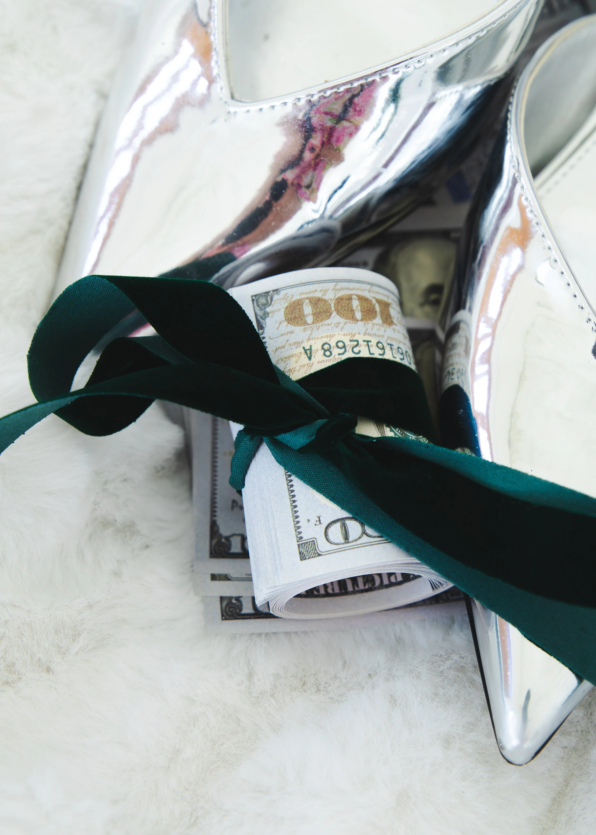 A silver metallic container holding rolled U.S. dollar bills and a black and green ribbon, placed on a white fuzzy surface.