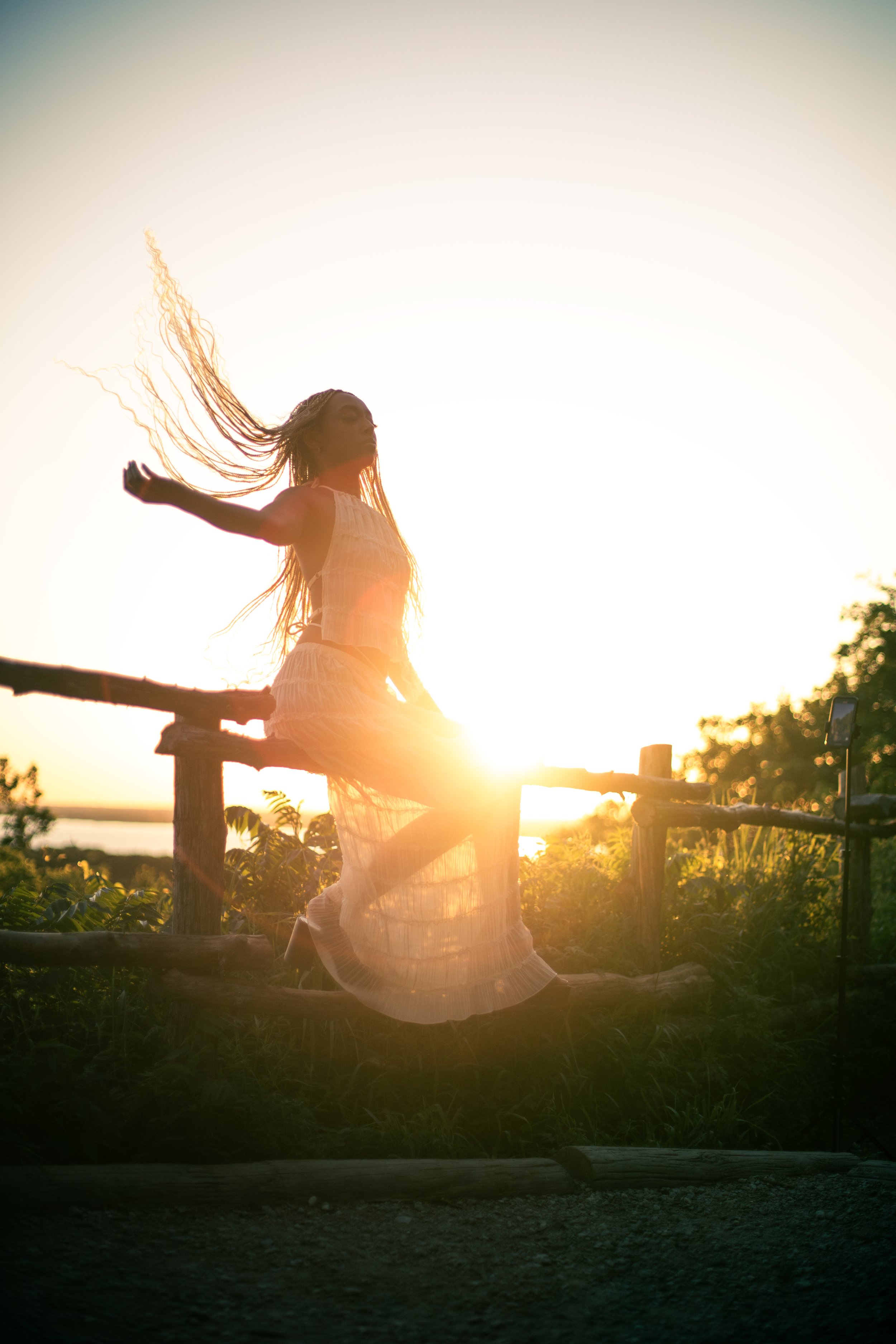 A woman with long hair standing with arms spread wide on a wooden fence at sunset, with sunlight illuminating her and the surrounding greenery.
