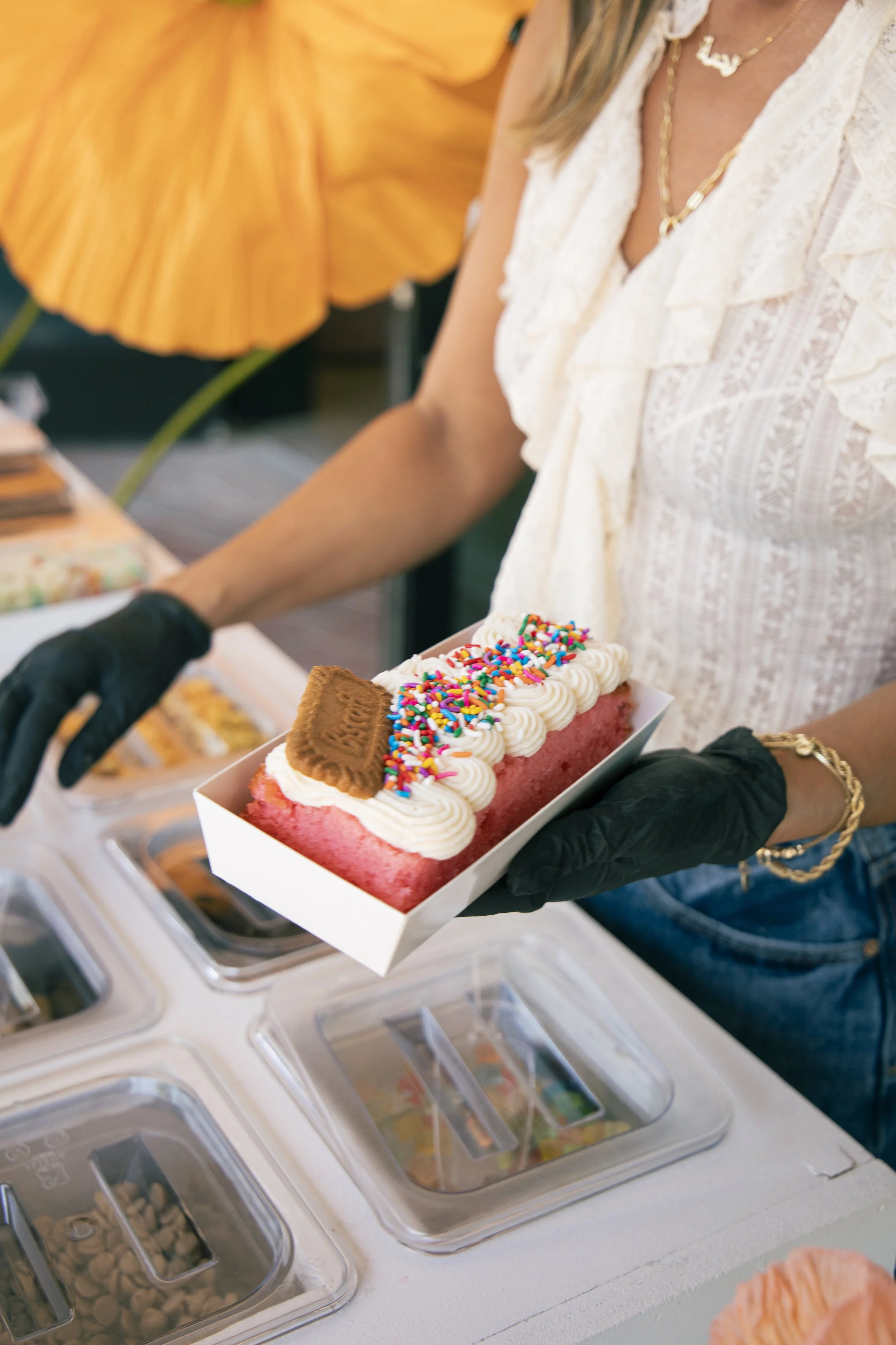 A woman is holding a rectangular cake with white frosting, colorful sprinkles, and a chocolate cookie on top. She is wearing black gloves and a white lace top. The background features a bright yellow parasol and several small containers of toppings o