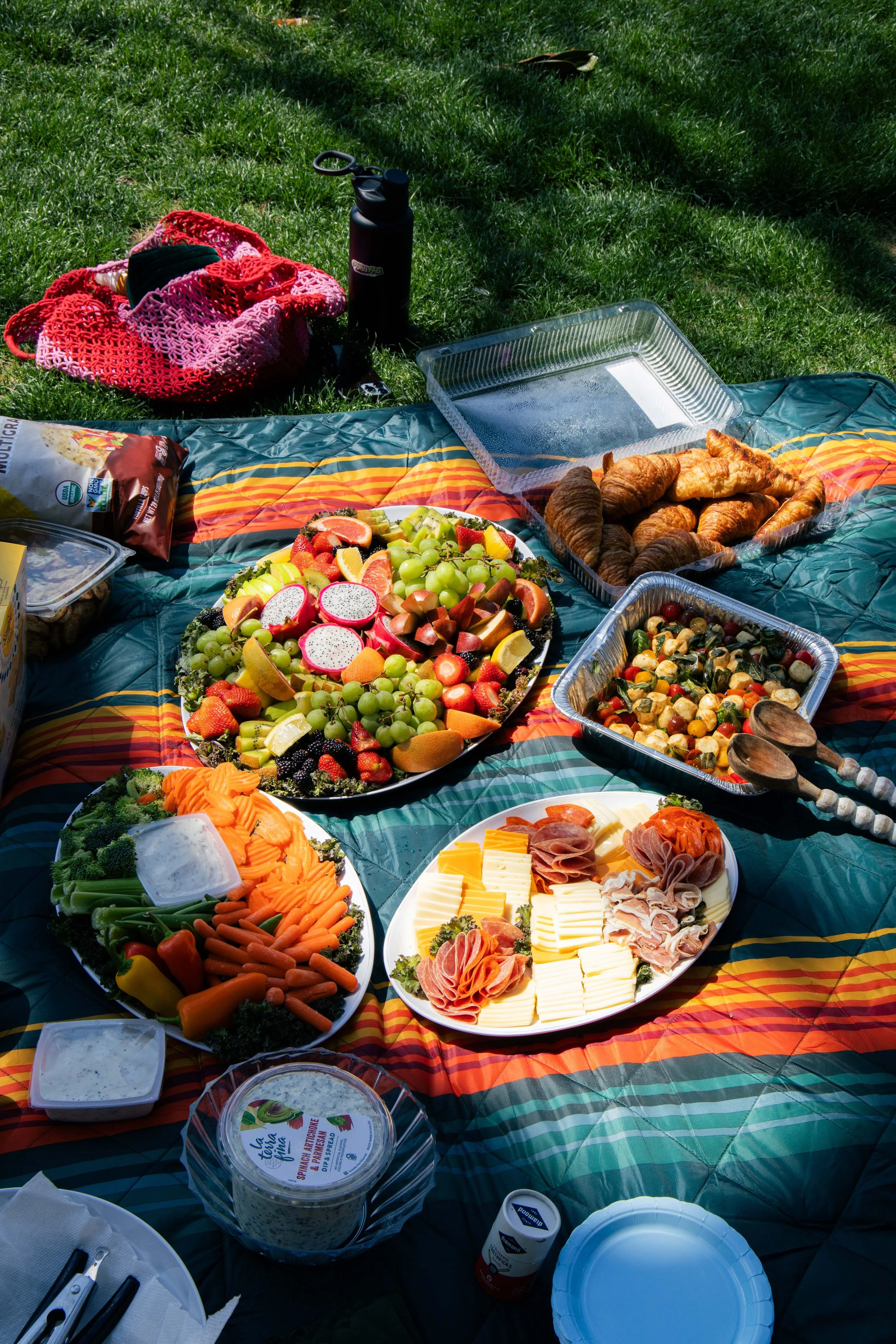 A picnic setup on a colorful blanket with a variety of food including a large fruit platter, croissants, sliced meat and cheese, vegetables, and small containers of sauces. There are also bottles and a pink crocheted bag on the grass.