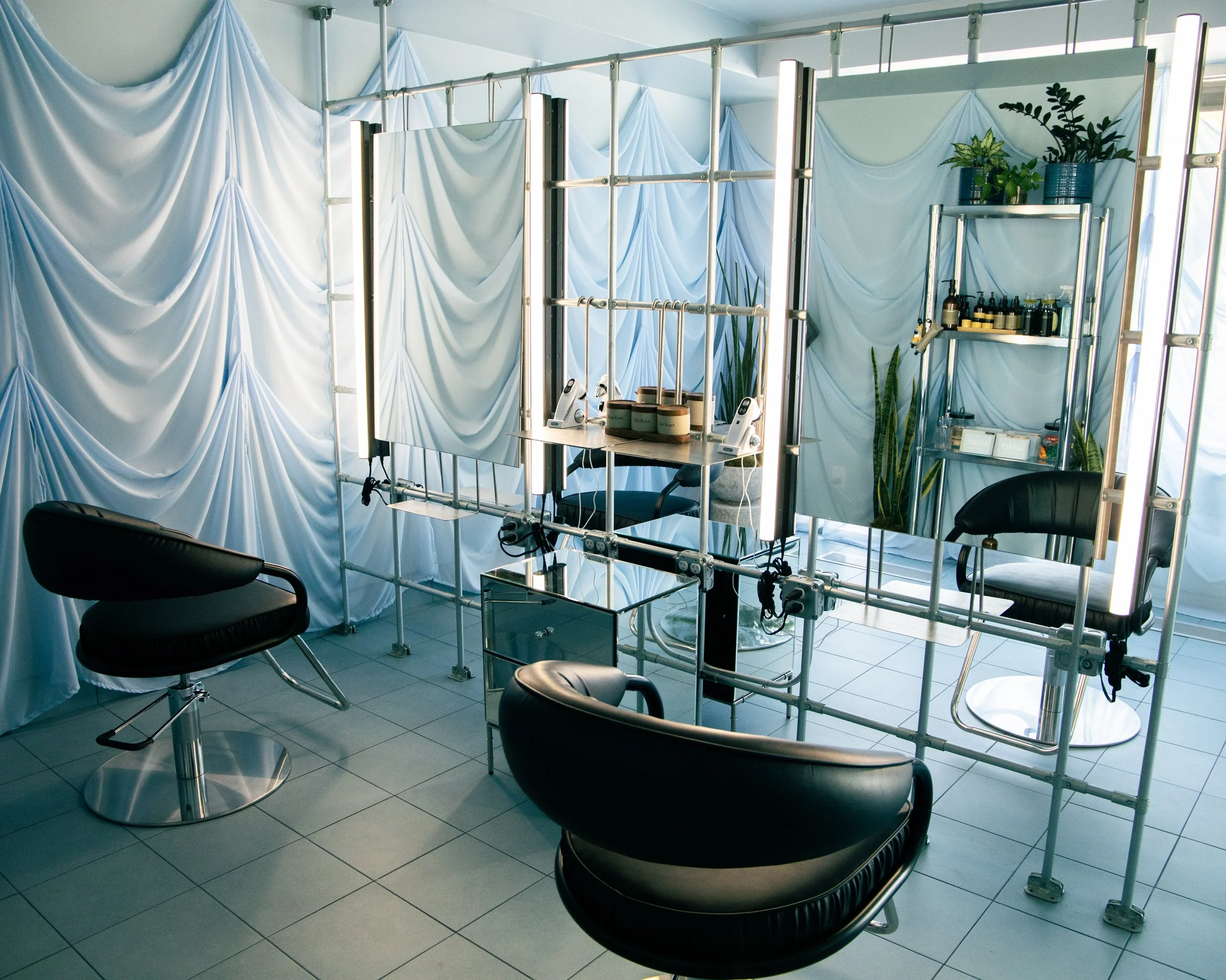 Interior of a beauty salon with two styling chairs, a mirror, and a shelf with hair products, against a backdrop of light blue draped curtains and a metal frame partition with potted plants.