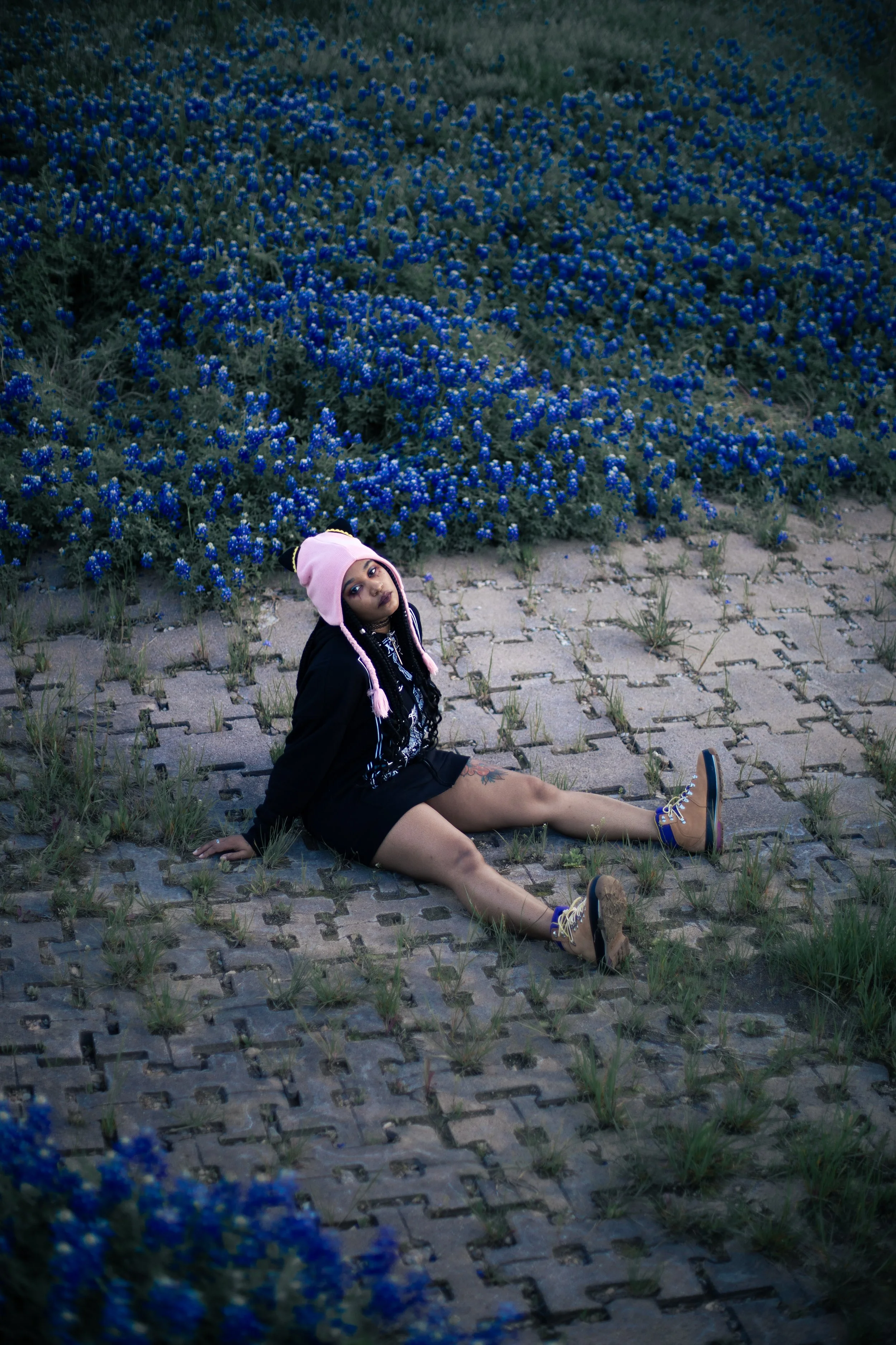 Local Dallas, Texas, DFW Non-traditional Fashion Editorial Creative Portraits Cover Art Photographer Photoshoot. Musician Igimeji . A young woman in a pink animal hat, black jacket, and shorts sitting on a paved area with grass, in front of a large b