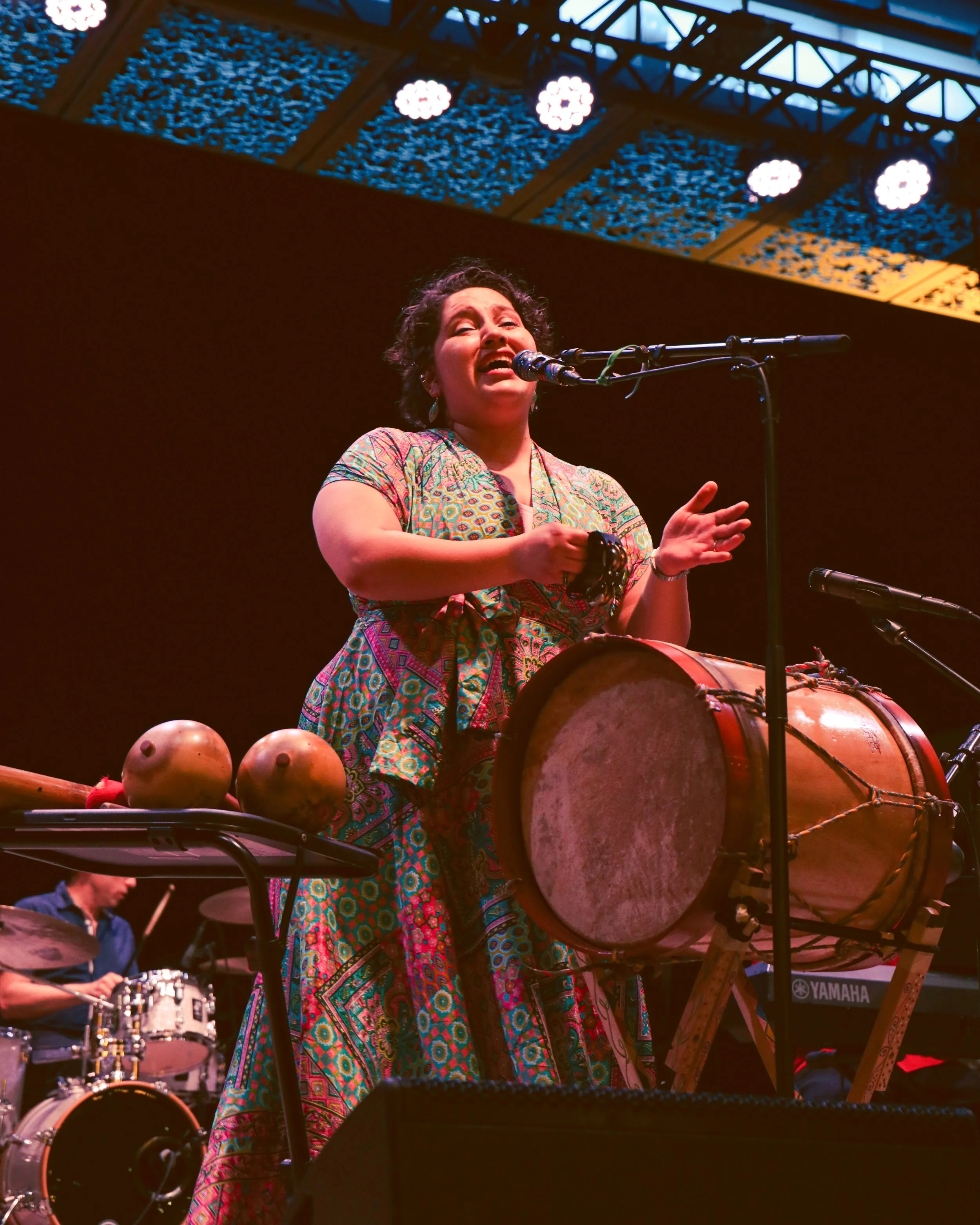 A woman singing into a microphone on stage, wearing a colorful patterned dress, with drums and a drummer visible in the background.