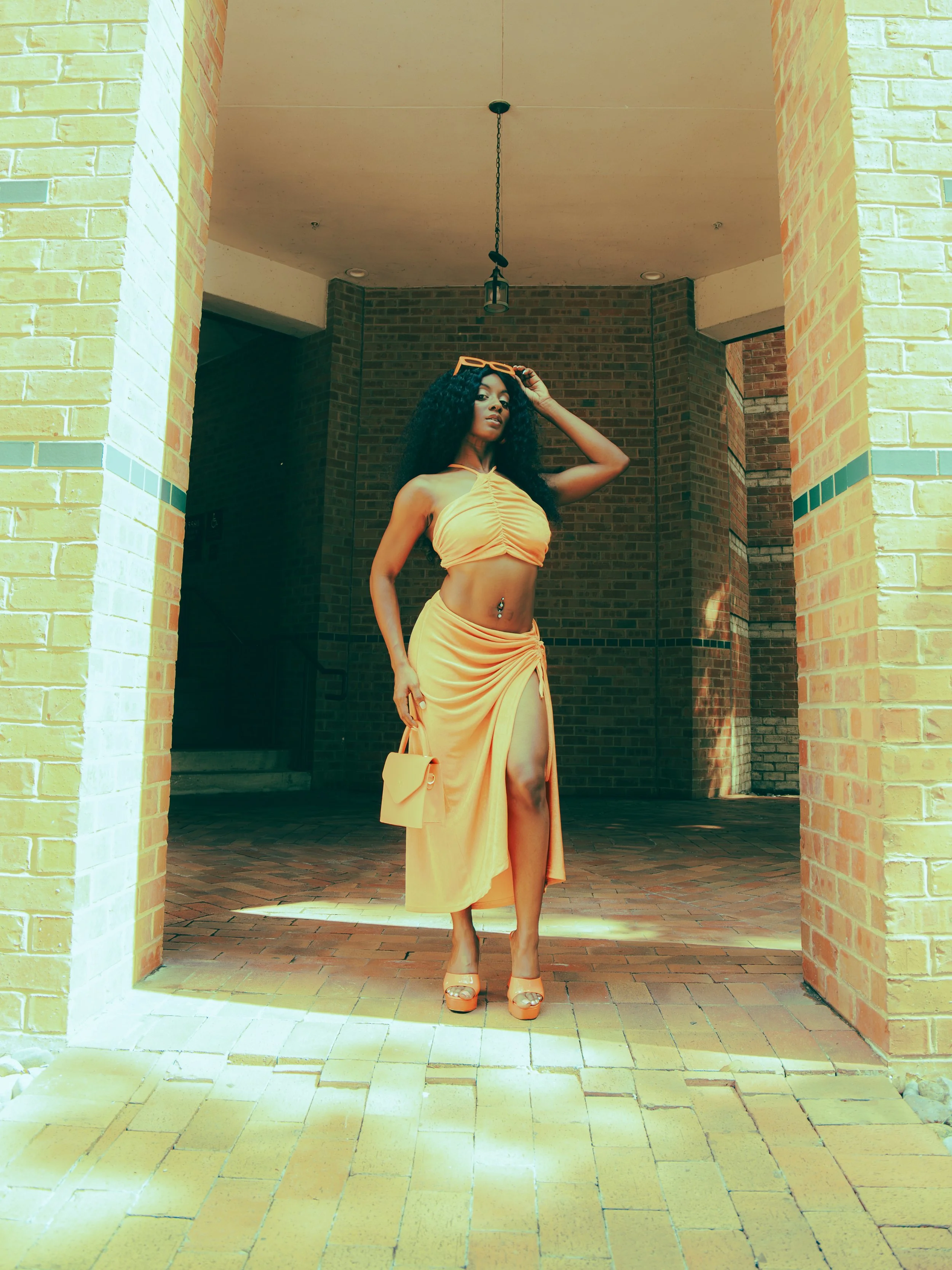 Local Dallas, Texas, DFW Non-traditional Fashion Editorial Creative Portraits Cover Art Photographer Photoshoot. A woman with long curly hair wearing a yellow bandeau top and matching long skirt with high slit, standing in an archway with brick walls