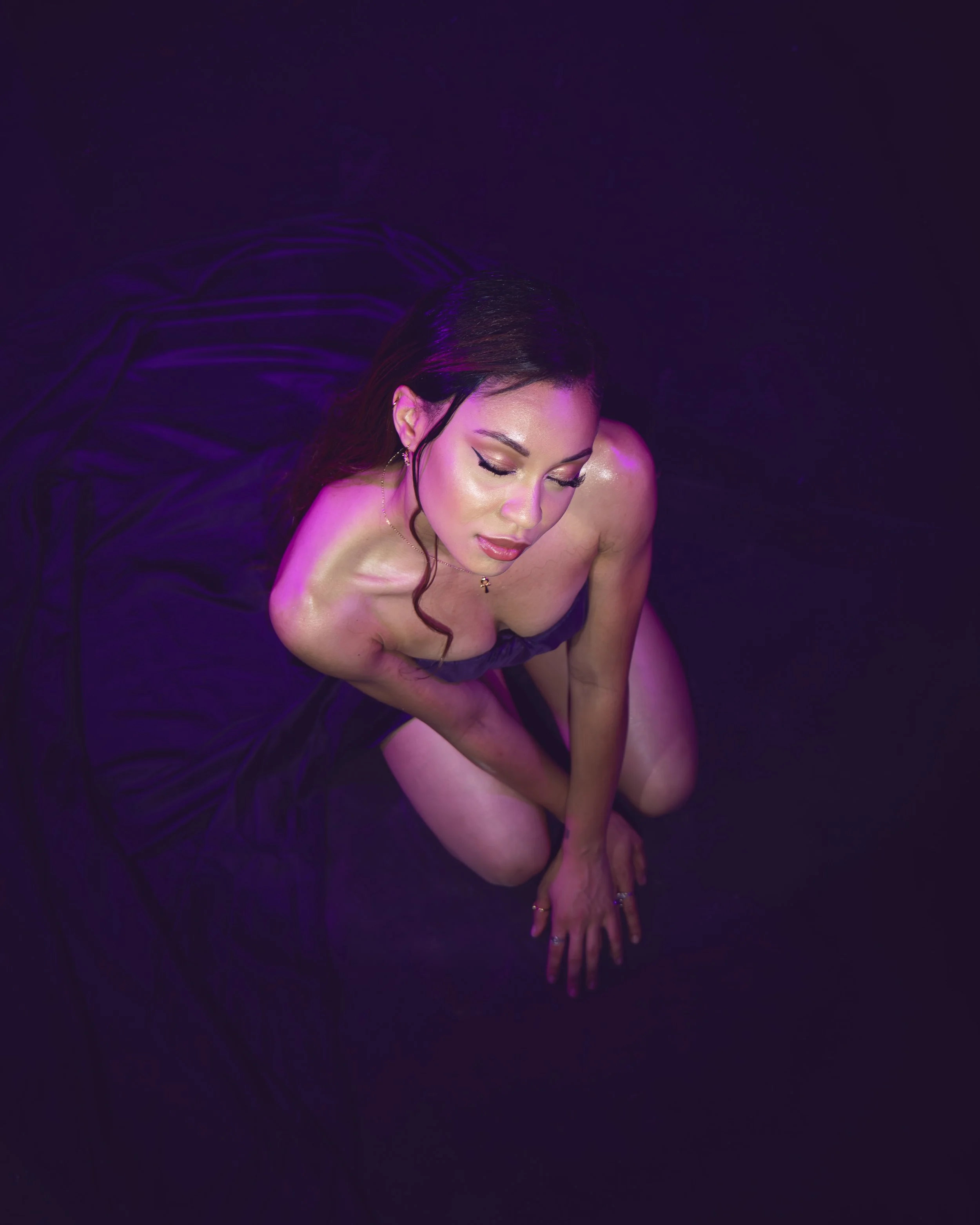 Local Dallas, Texas, DFW Non-traditional Fashion Editorial Creative Portraits Cover Art Photographer Photoshoot. Musician Rebelle Flor . A woman with dark hair and makeup, wearing earrings and a necklace, poses in a dark environment with purple light