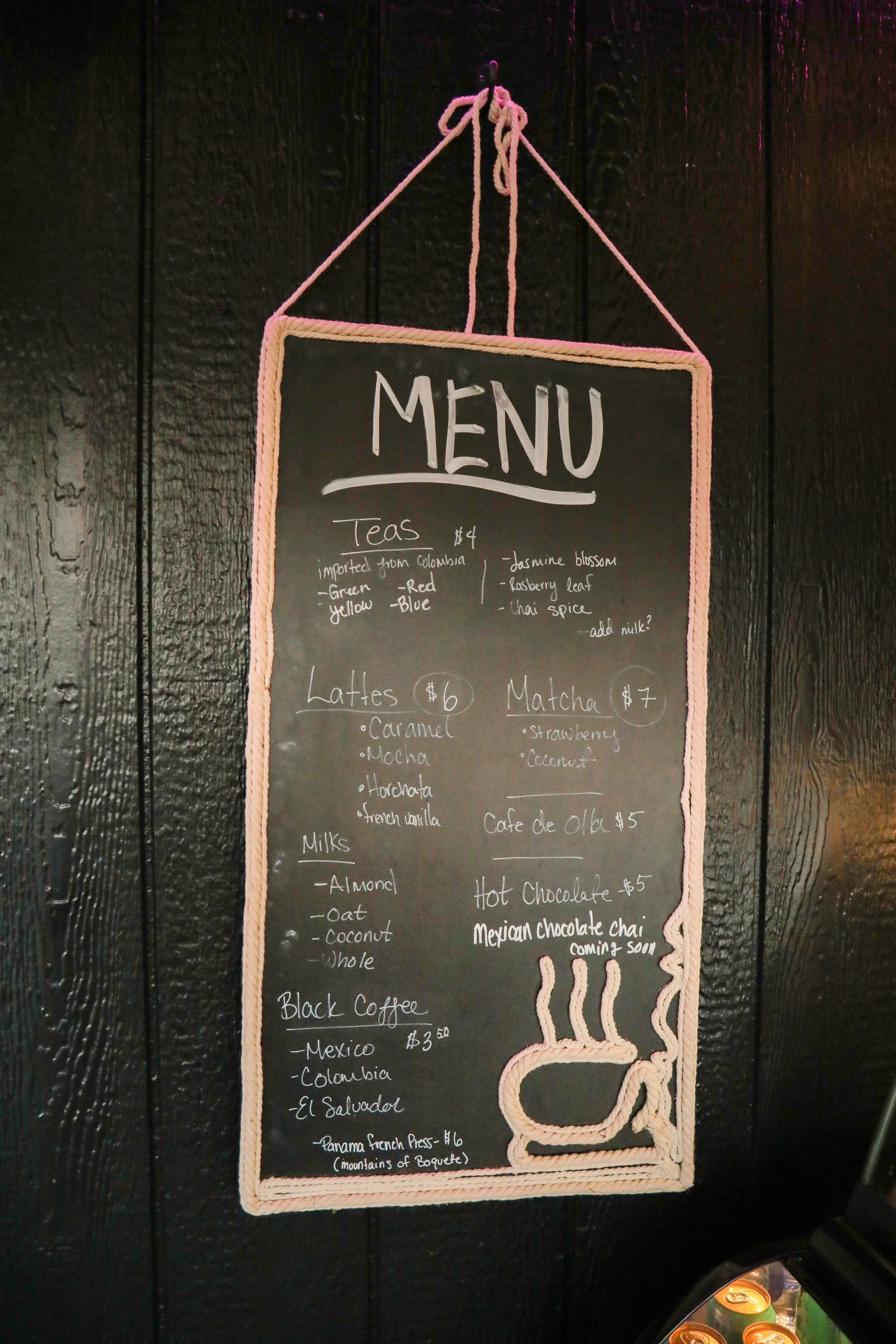 Black chalkboard menu with handwritten tea, latte, and hot chocolate options, hanging on a black wall with pink decorative border.