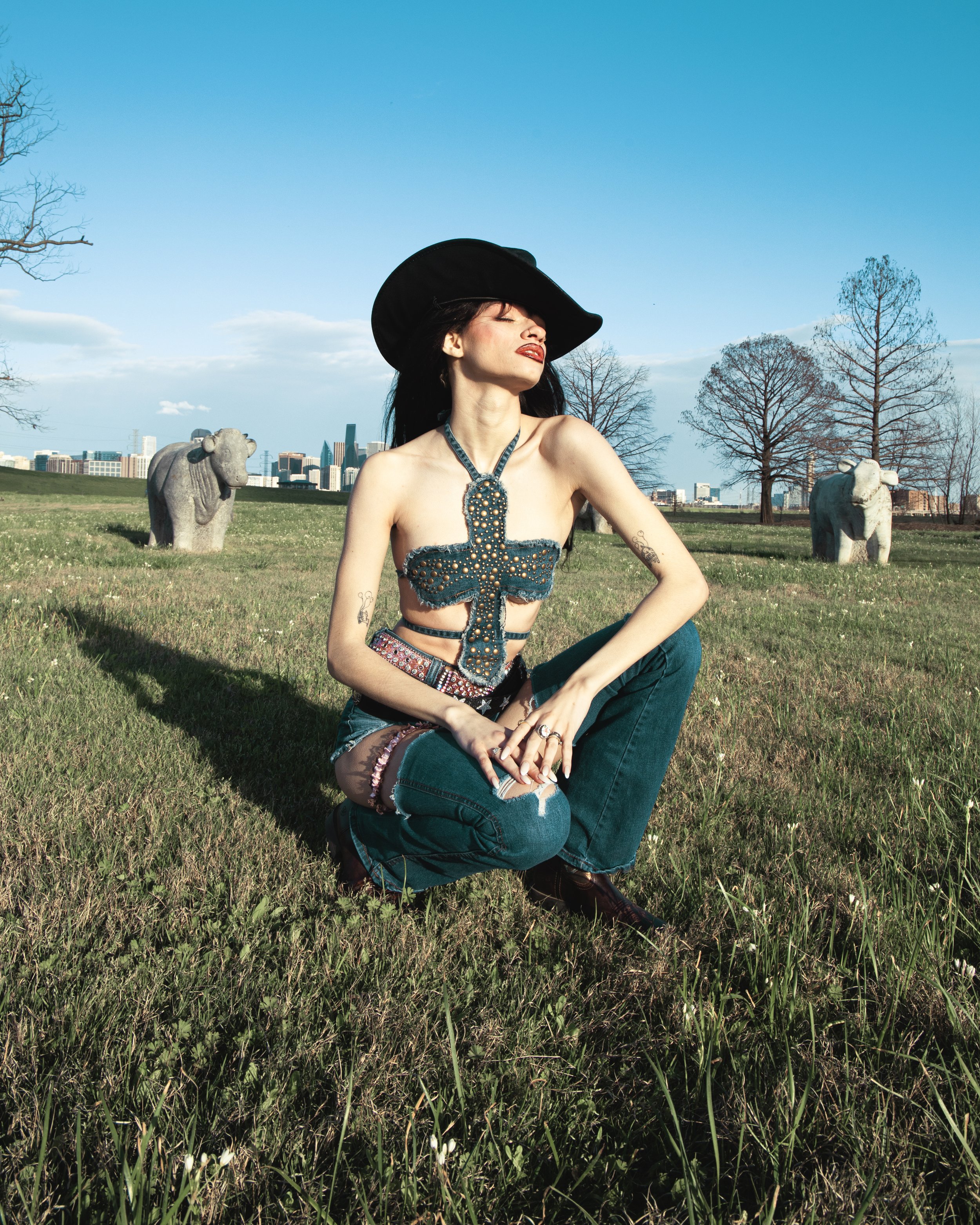 Local Dallas, Texas, DFW Non-traditional Fashion Editorial Creative Portraits Cover Art Photographer Photoshoot. Trammell Crow Park . A woman with tattoos on her arms is sitting on grass in a park, wearing a black hat and a denim top with pearl embel