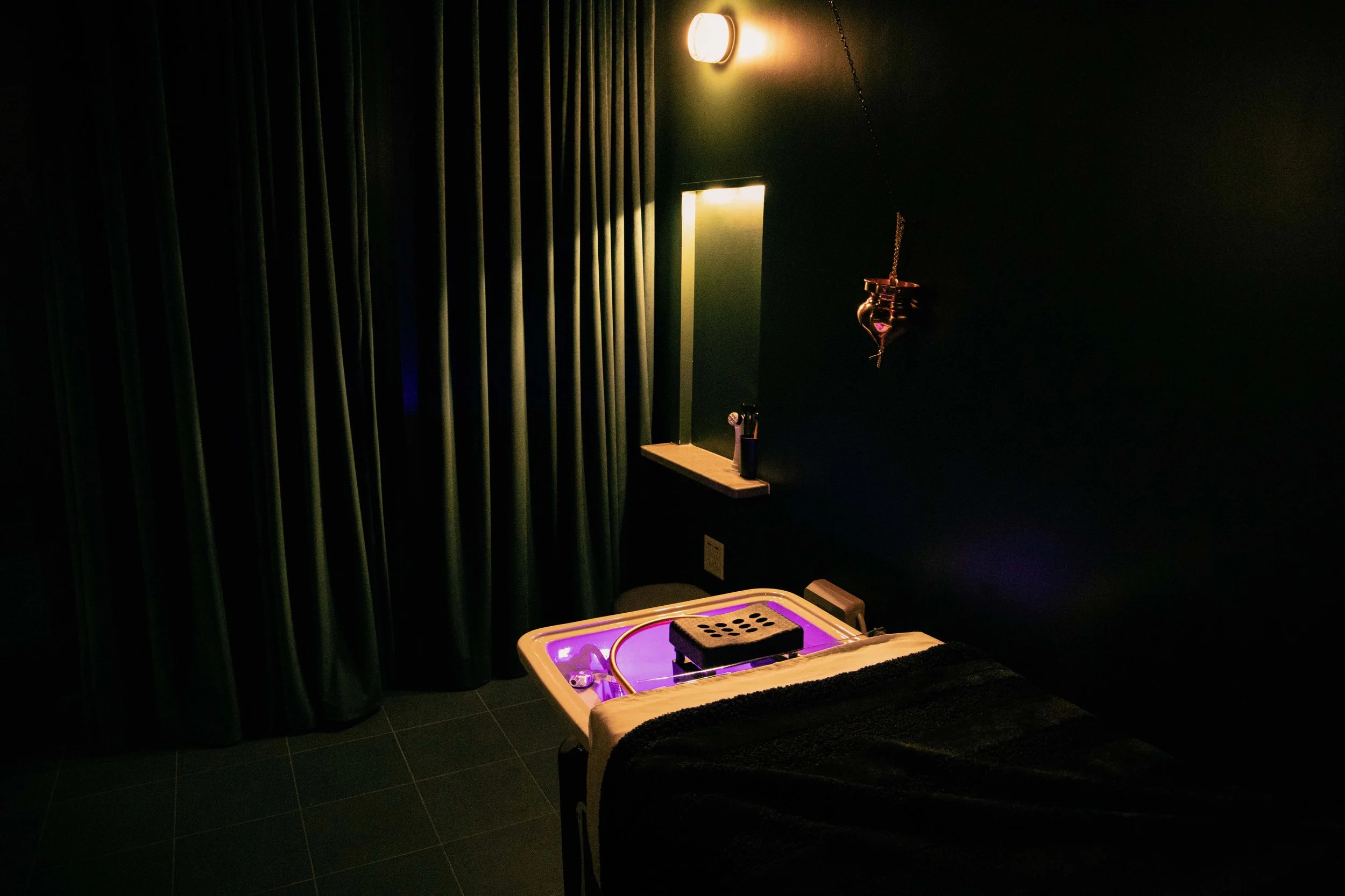 A small room with dark walls and tiled floor, illuminated by warm yellow lighting from a wall-mounted fixture. On the right side, there is a wooden table with electronic equipment and purple lighting underneath. A green curtain covers the left side o