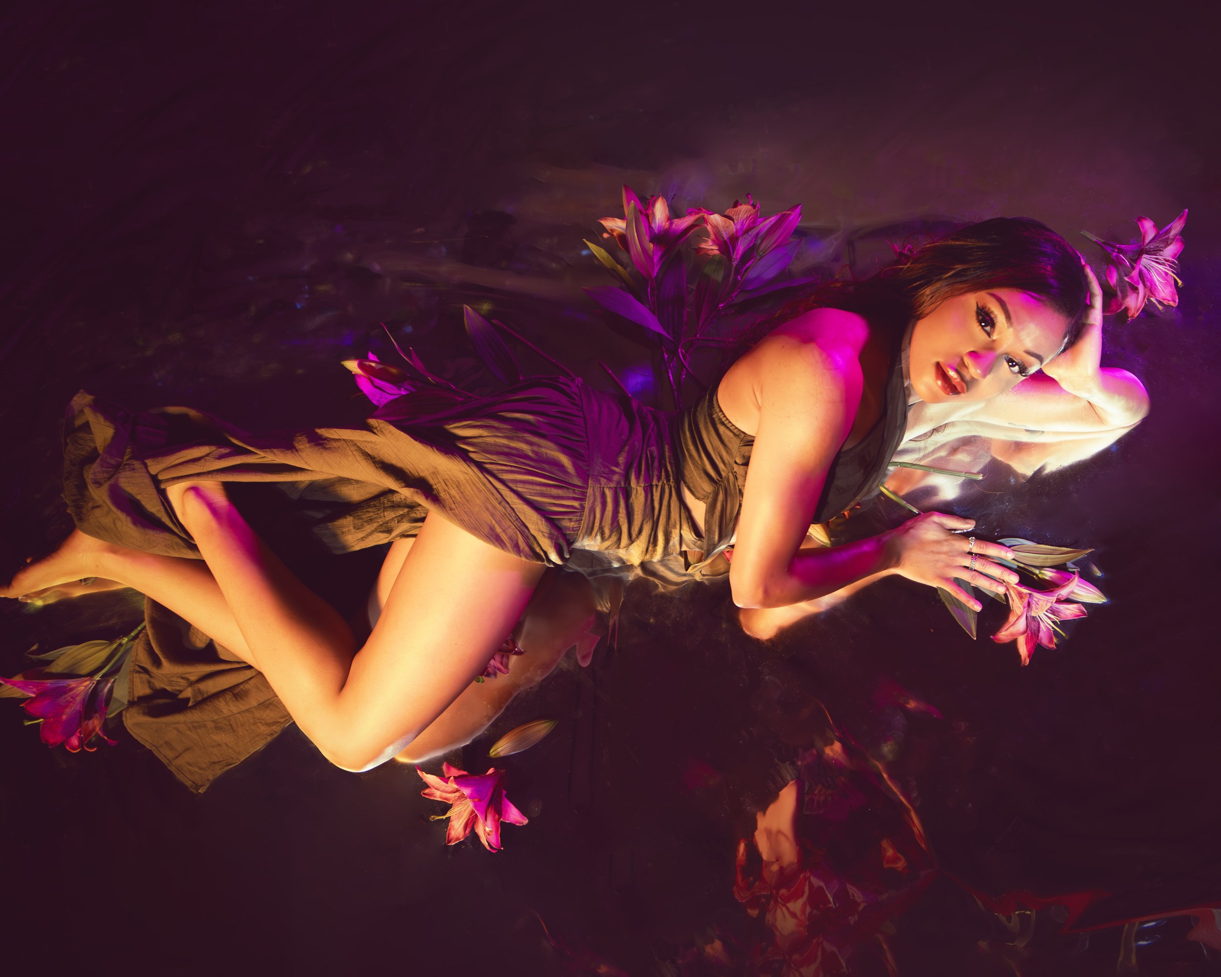 Local Dallas, Texas, DFW Non-traditional Fashion Editorial Creative Portraits Cover Art Photographer Photoshoot. Musician Rebelle Flor. A woman with dark hair and makeup lying on her side in water illuminated with pink and purple lighting, surrounded