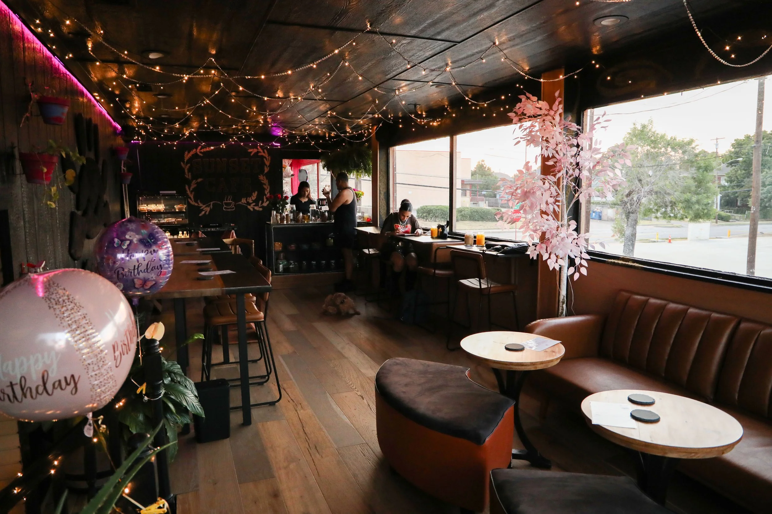 A cozy cafe interior decorated for a birthday celebration with balloons, fairy lights, and pink foliage outside the large windows. Several people are sitting and standing inside, and a dog lies on the wooden floor.