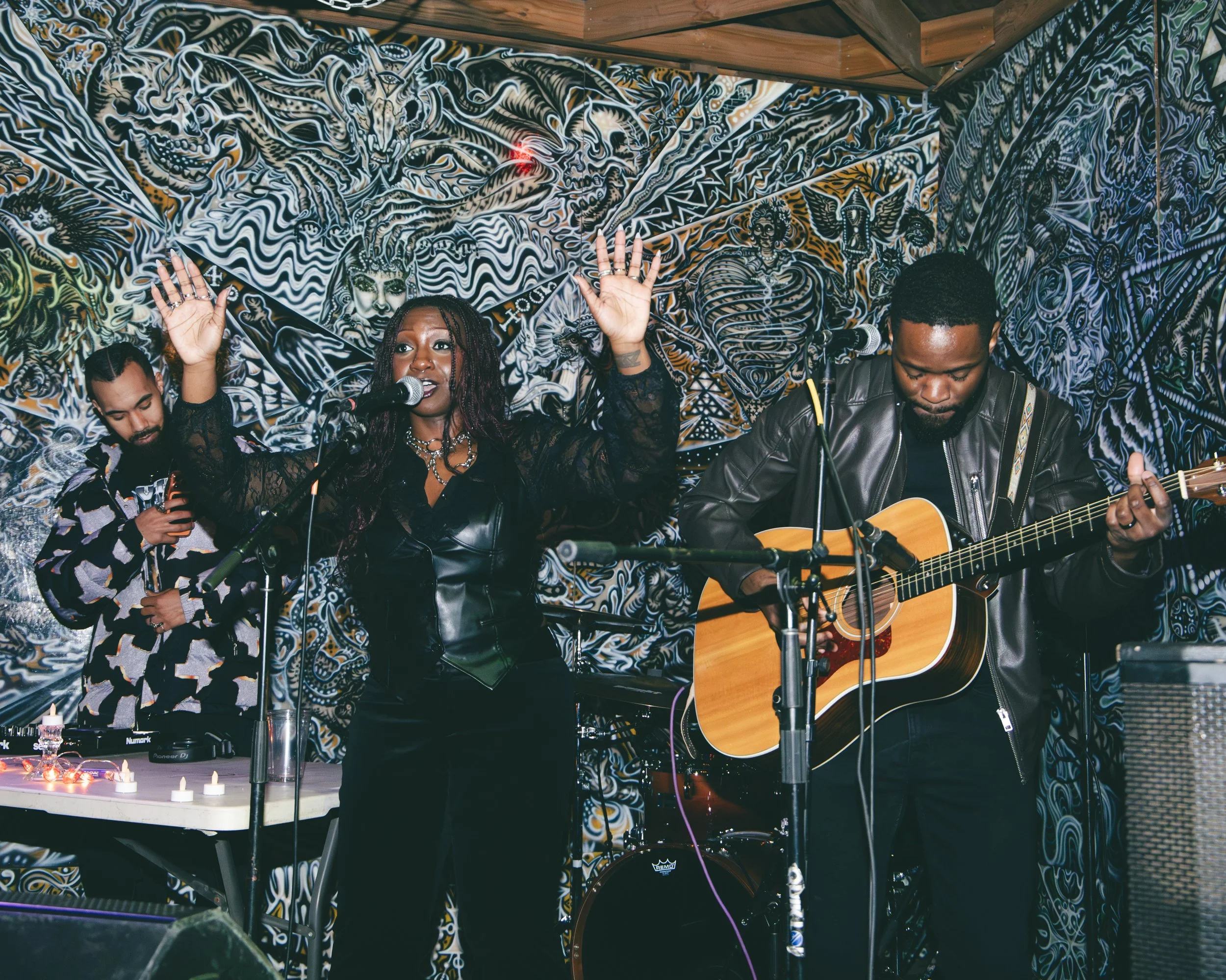 Three musicians performing in front of a psychedelic-black-and-white mural. The woman in the center has her eyes closed, with braids, and is singing into a microphone with her hands raised. The man to her right plays an acoustic guitar, and the man o