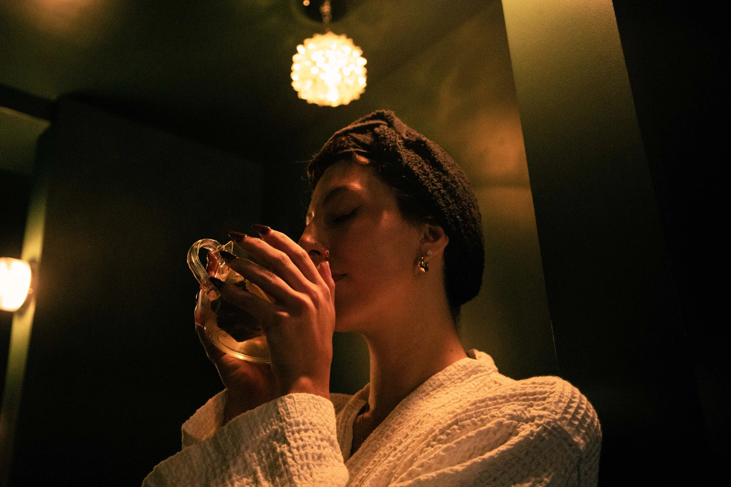 A woman with dark hair wearing a towel keeps her eyes closed while drinking from a glass at night in a dimly lit room