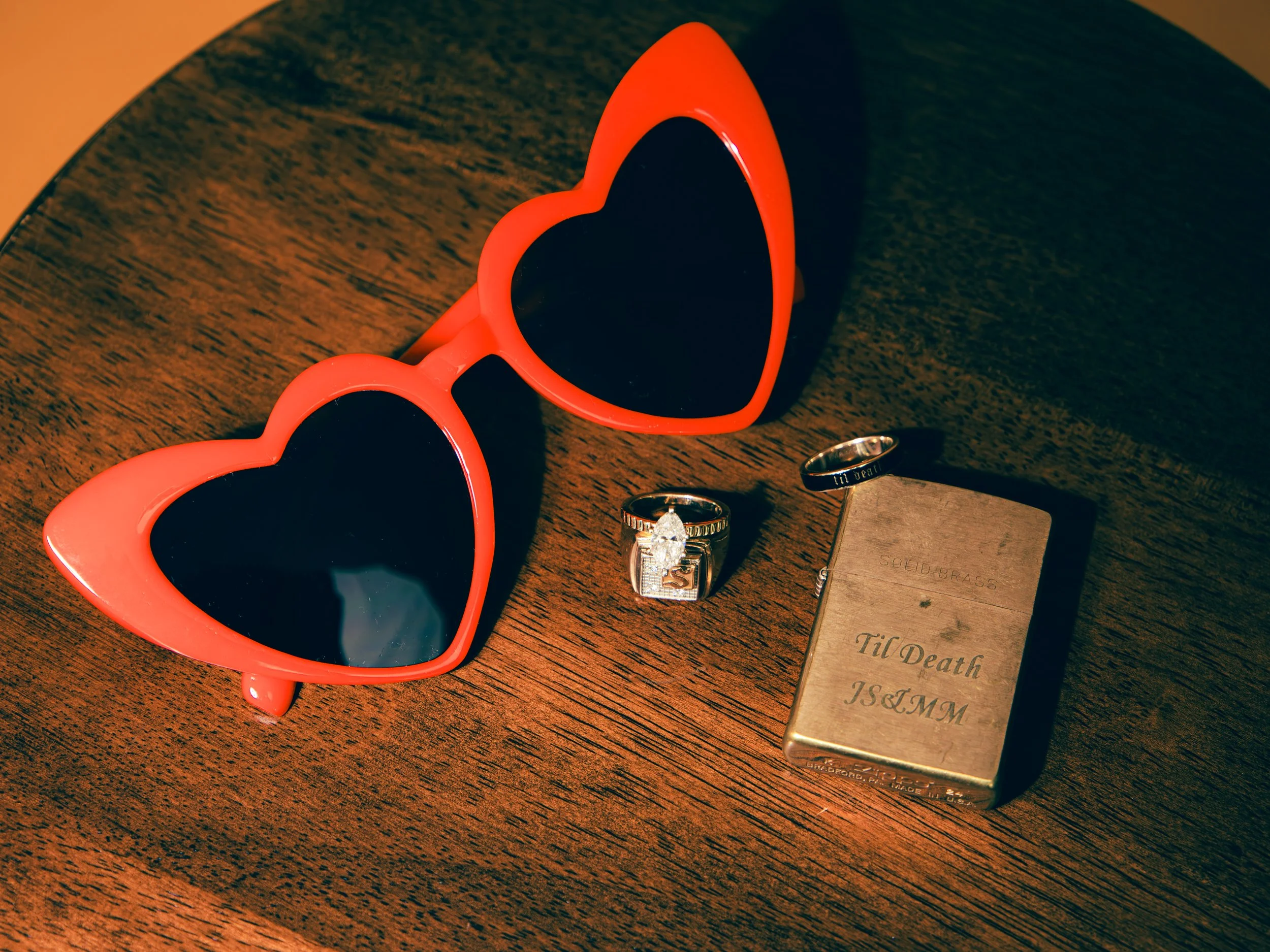 A pair of red, heart-shaped sunglasses on a wooden surface. Nearby are two silver rings, one of which has a prominent large clear gemstone, and a wooden box with engraved text.