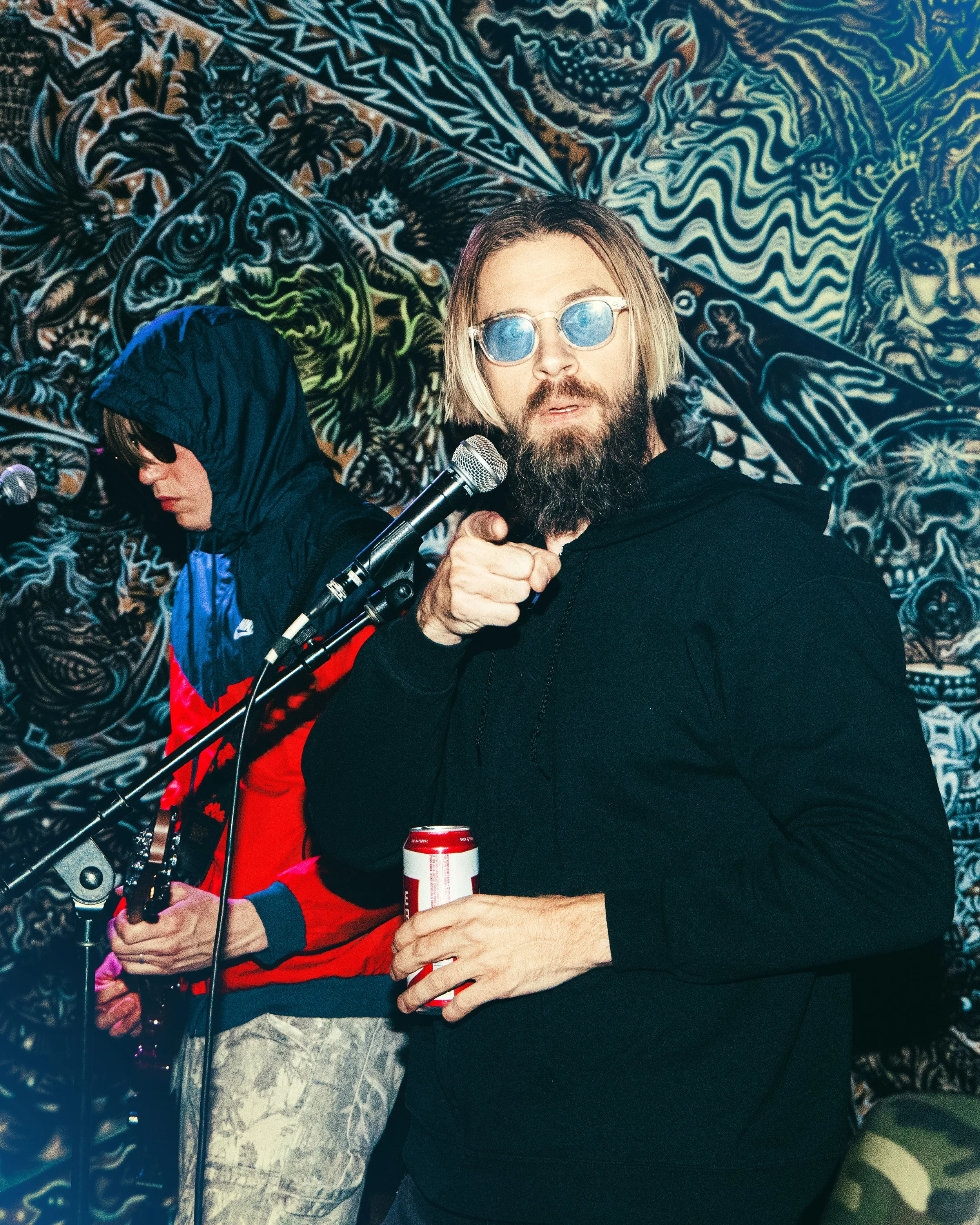 A man with long hair and a beard wearing reflective sunglasses, holding a can of beer, standing in front of a vibrant, colorful mural featuring various abstract and mythological creatures.