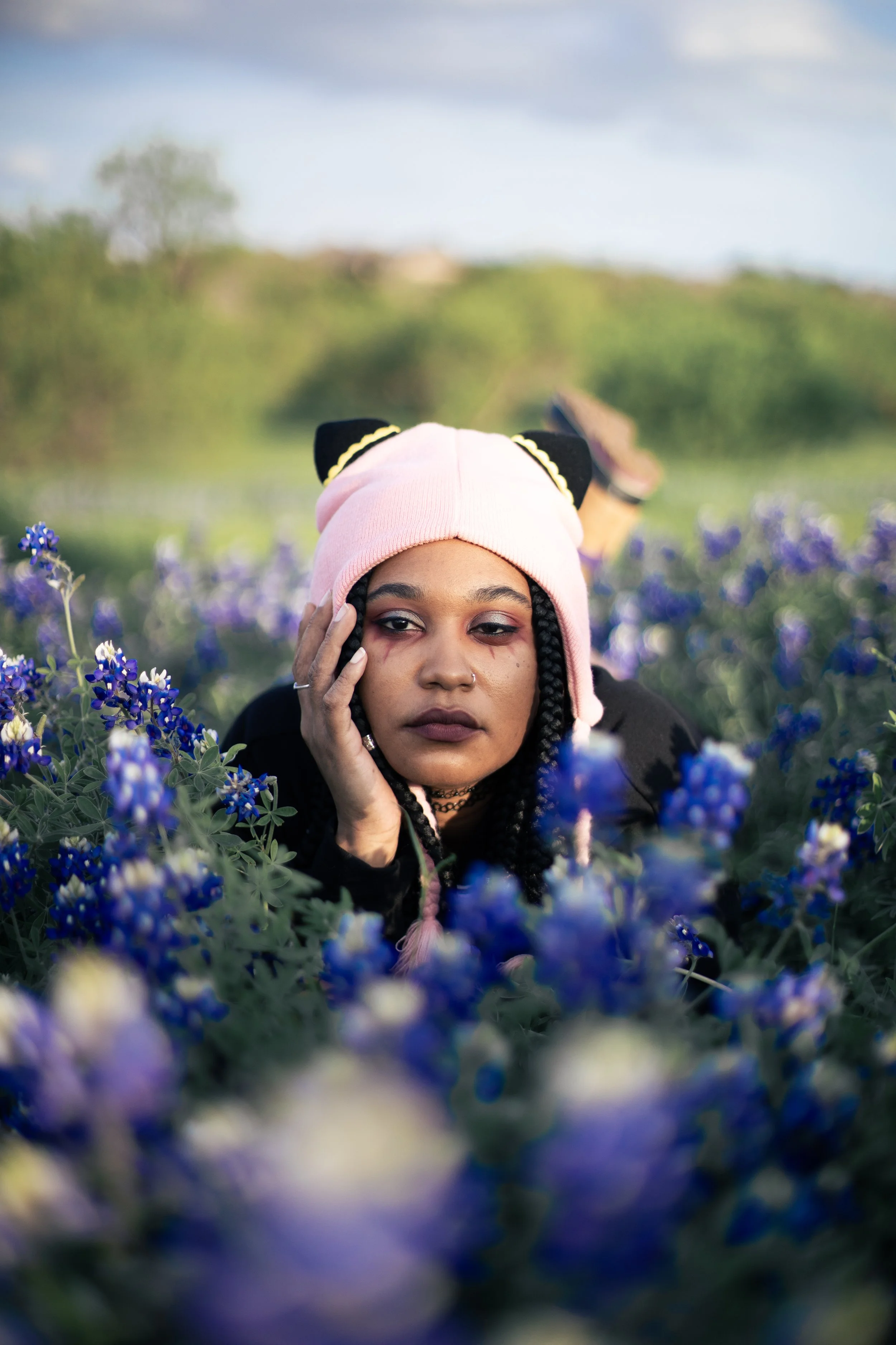 Local Dallas, Texas, DFW Non-traditional Fashion Editorial Creative Portraits Cover Art Photographer Photoshoot. Musician Igimeji . A young woman with braided hair, wearing a pink nightcap with black cat ears, lying in a field of purple flowers, with