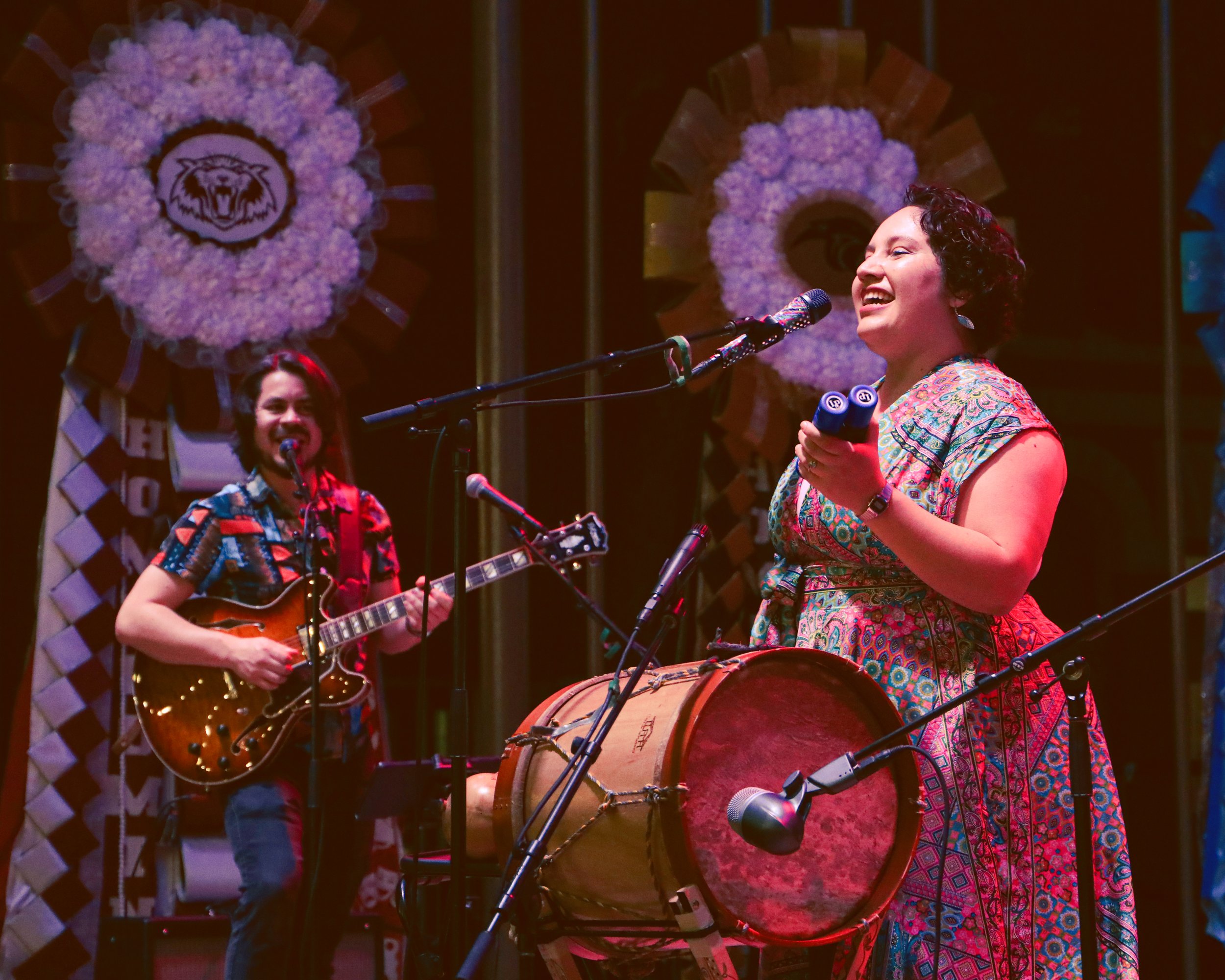 Two musicians performing on stage, one playing guitar and the other singing into a microphone. The background features decorative floral wreaths with a tiger emblem. The performers are dressed in colorful clothing.