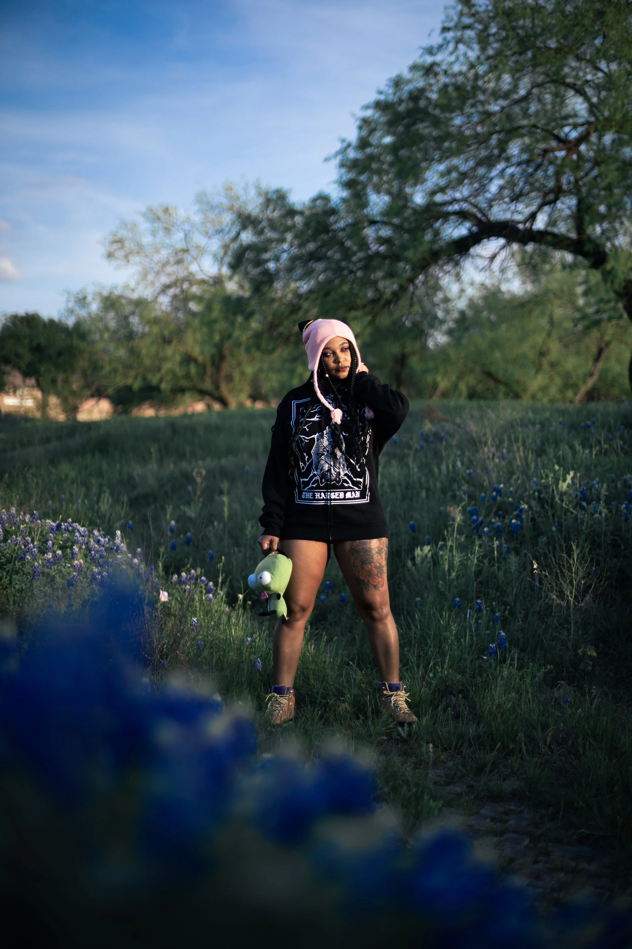  Local Dallas, Texas, DFW Non-traditional Fashion Editorial Creative Portraits Cover Art Photographer Photoshoot. Musician Igimeji .A woman standing in a grassy field with purple flowers, wearing a pink beanie, a black hoodie, and boots, holding a gr