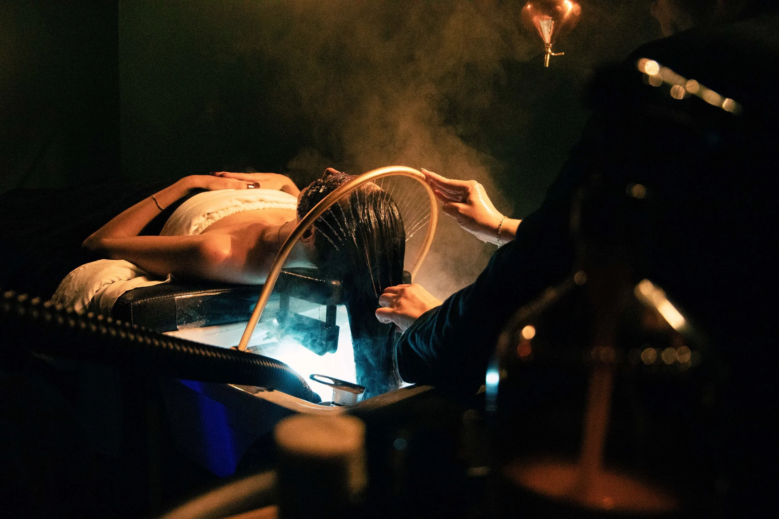 A person lying on a medical table with their head tilted back, receiving a treatment involving a hoop held by a healthcare professional, with smoke or vapor in the scene, in a dark room with orange lighting.