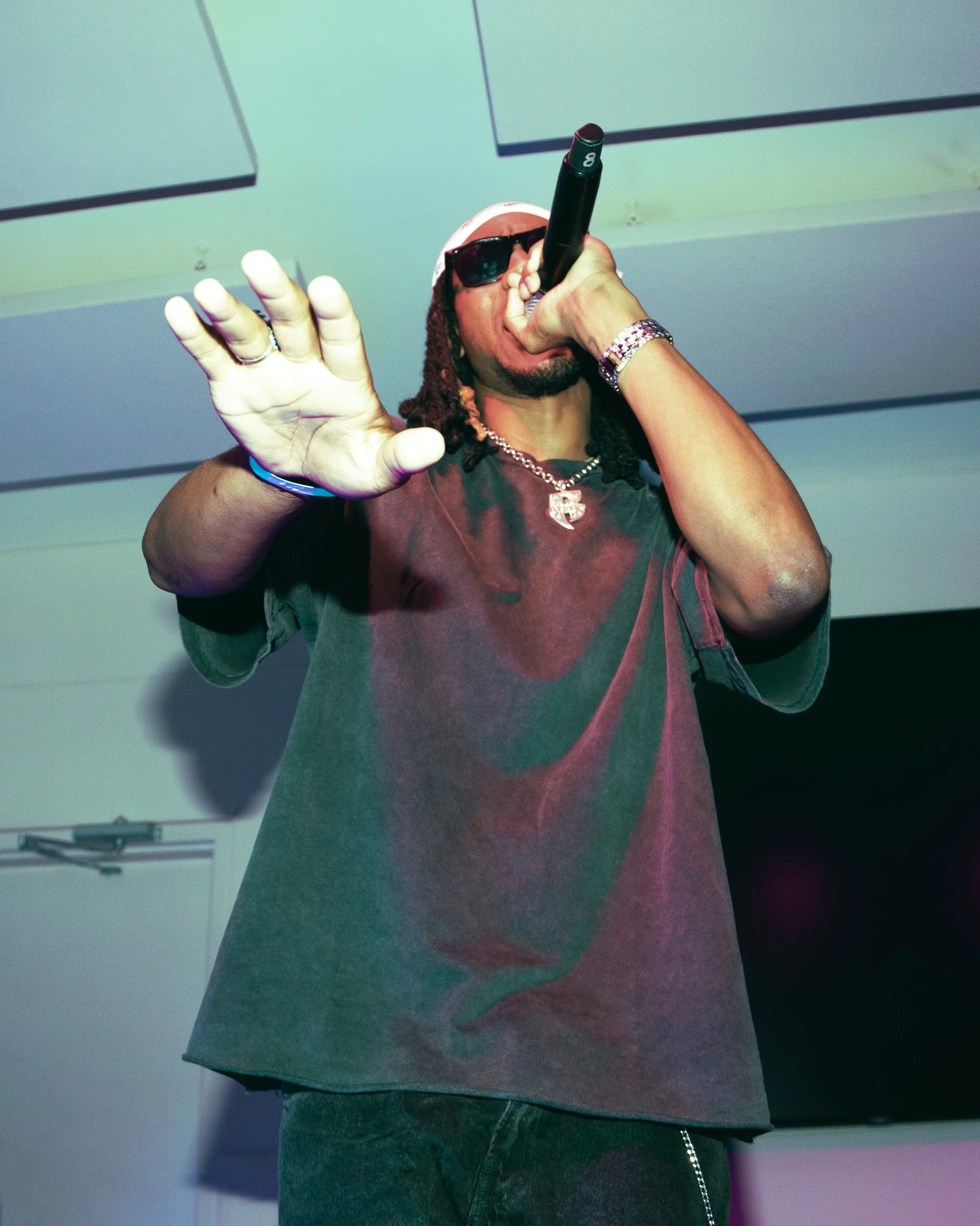 Person wearing sunglasses, a red cap, diamond jewelry, and a dark t-shirt, holding a microphone and performing on stage with a blue background.