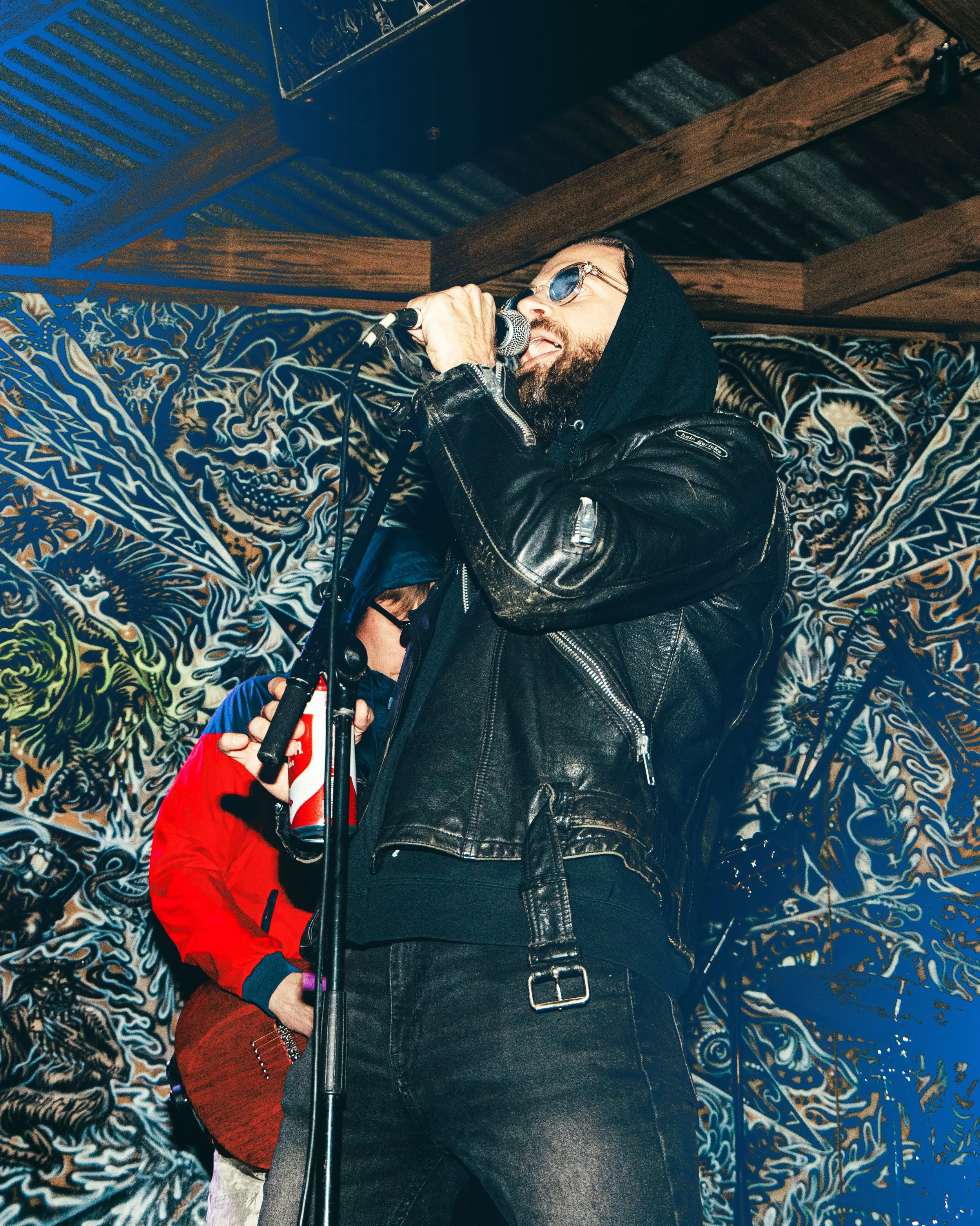 A man singing into a microphone, dressed in a black leather jacket and sunglasses, with a tattooed arm and a beard, standing against a colorful, artistic wall background.