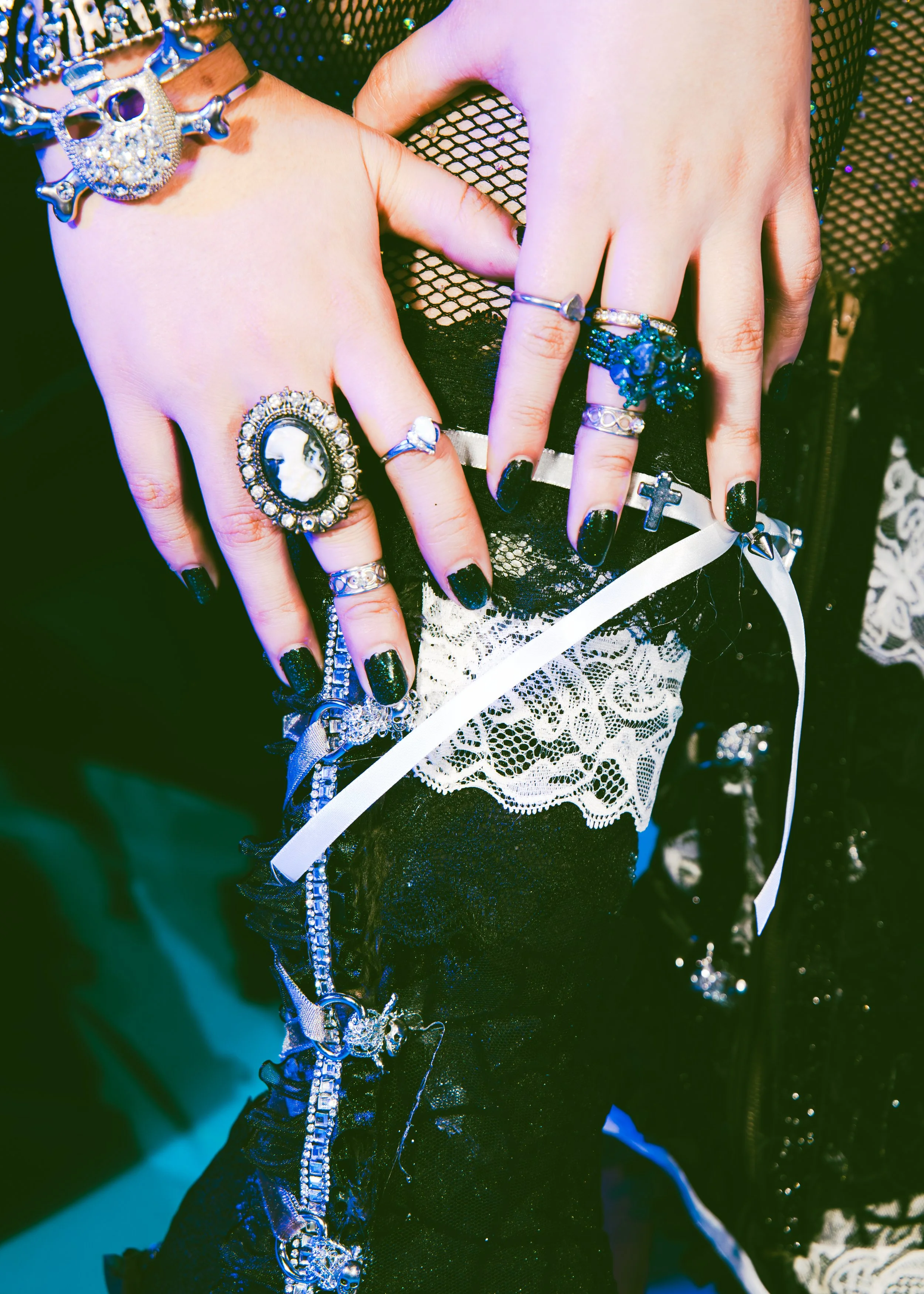 Close-up of a person's hands showing several decorative rings, a bracelet with skull and chain details, and black nail polish, with focus on jewelry and a fishnet sleeve against a dark background.