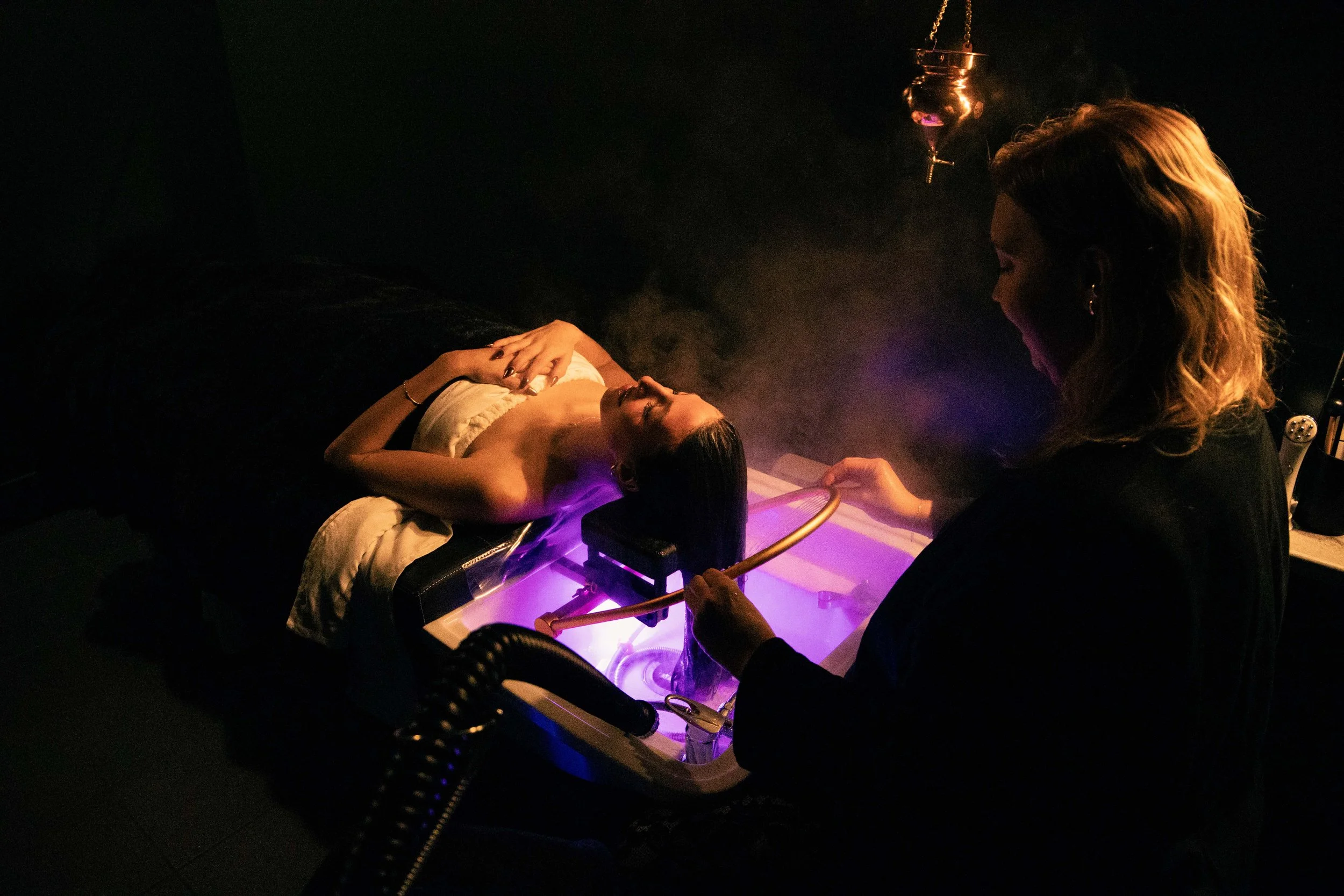 A woman lying on a medical table with her arm extended, receiving a treatment from a female practitioner in a dark room illuminated by purple lighting. The practitioner is using medical equipment near the woman's arm, possibly performing a laser or l