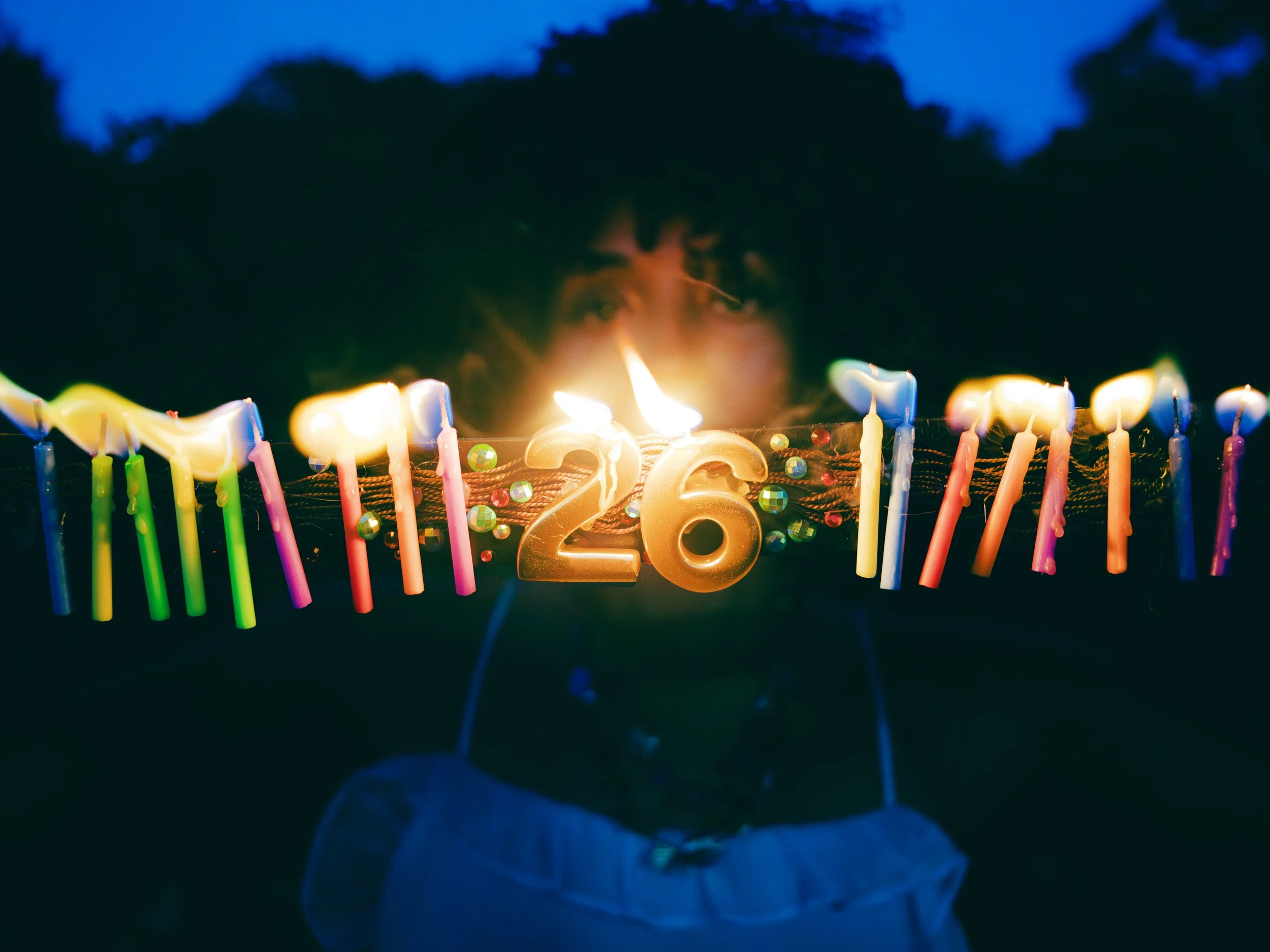 A birthday cake with lit candles and a golden number '26' candle, with a dark background and blue sky, celebrating a 26th birthday.