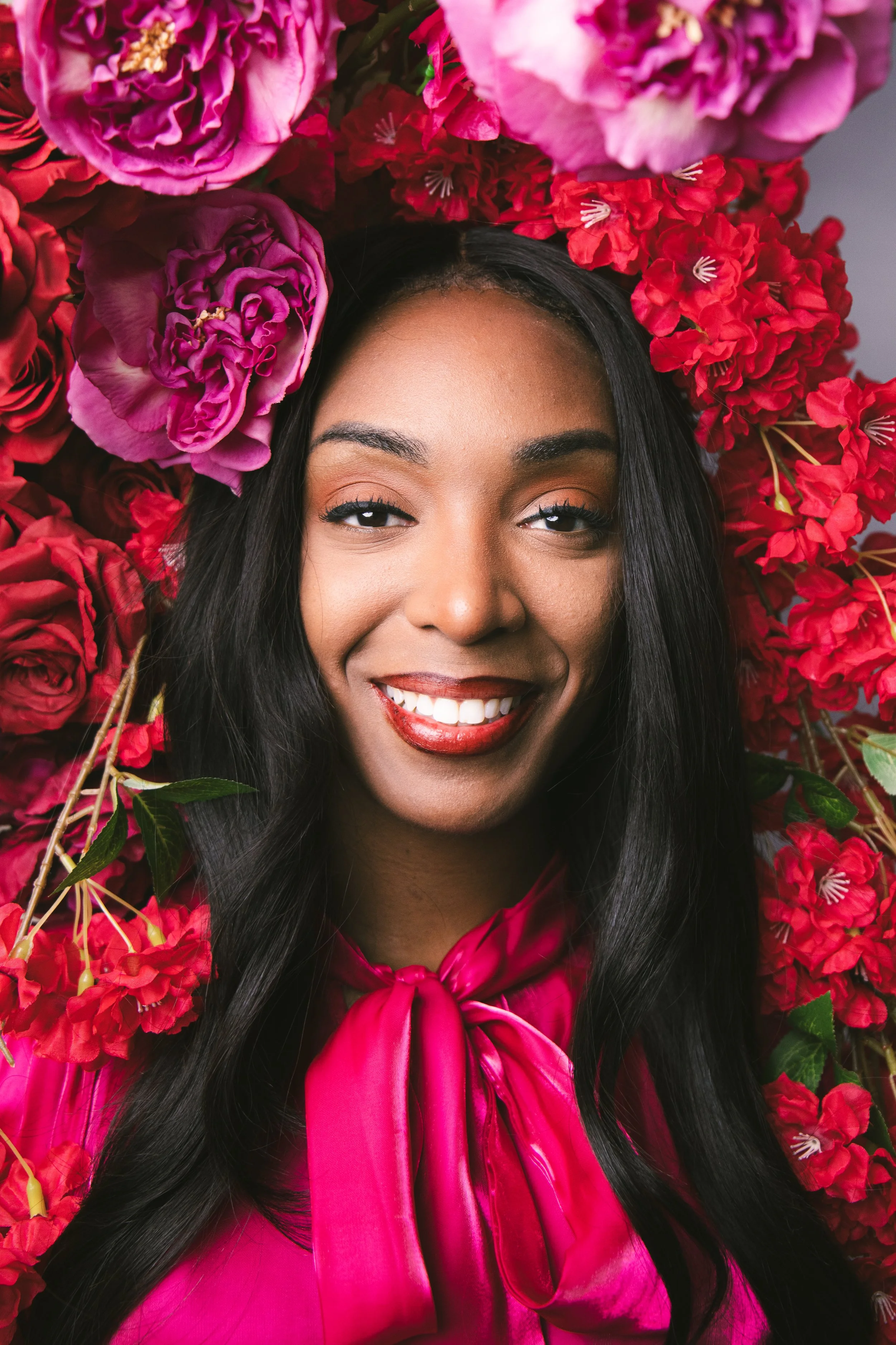 Local Dallas, Texas, DFW Non-traditional Fashion Editorial Creative Portraits Cover Art Photographer Photoshoot. A smiling woman with long black hair, wearing a pink shiny dress and surrounded by pink and red flowers.
