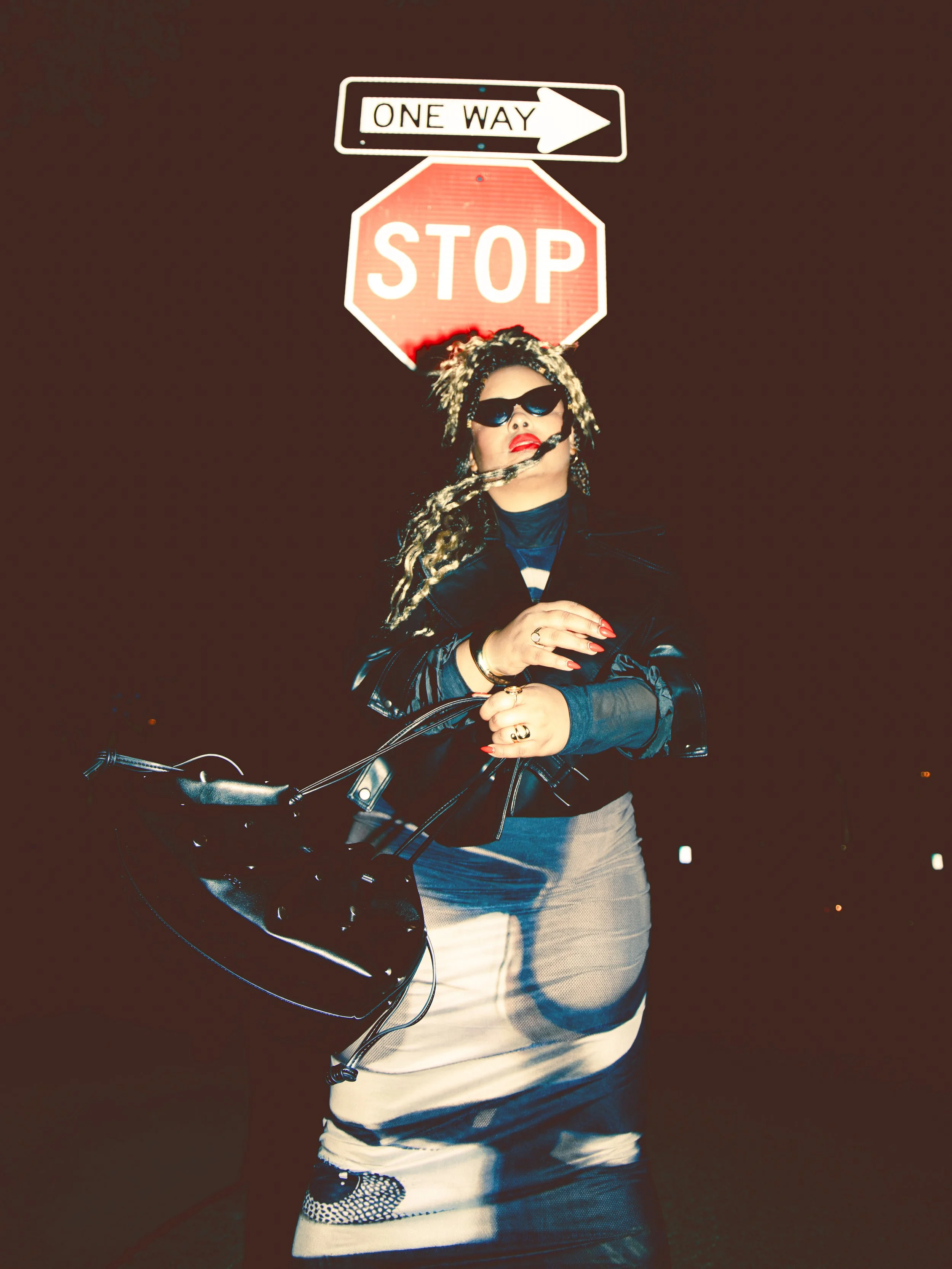 Local Dallas, Texas, DFW Non-traditional Fashion Editorial Creative Portraits Cover Art Photographer Photoshoot. A woman with blonde dreadlocks, wearing sunglasses and red lipstick, standing in front of a stop sign and one-way sign at night, holding 