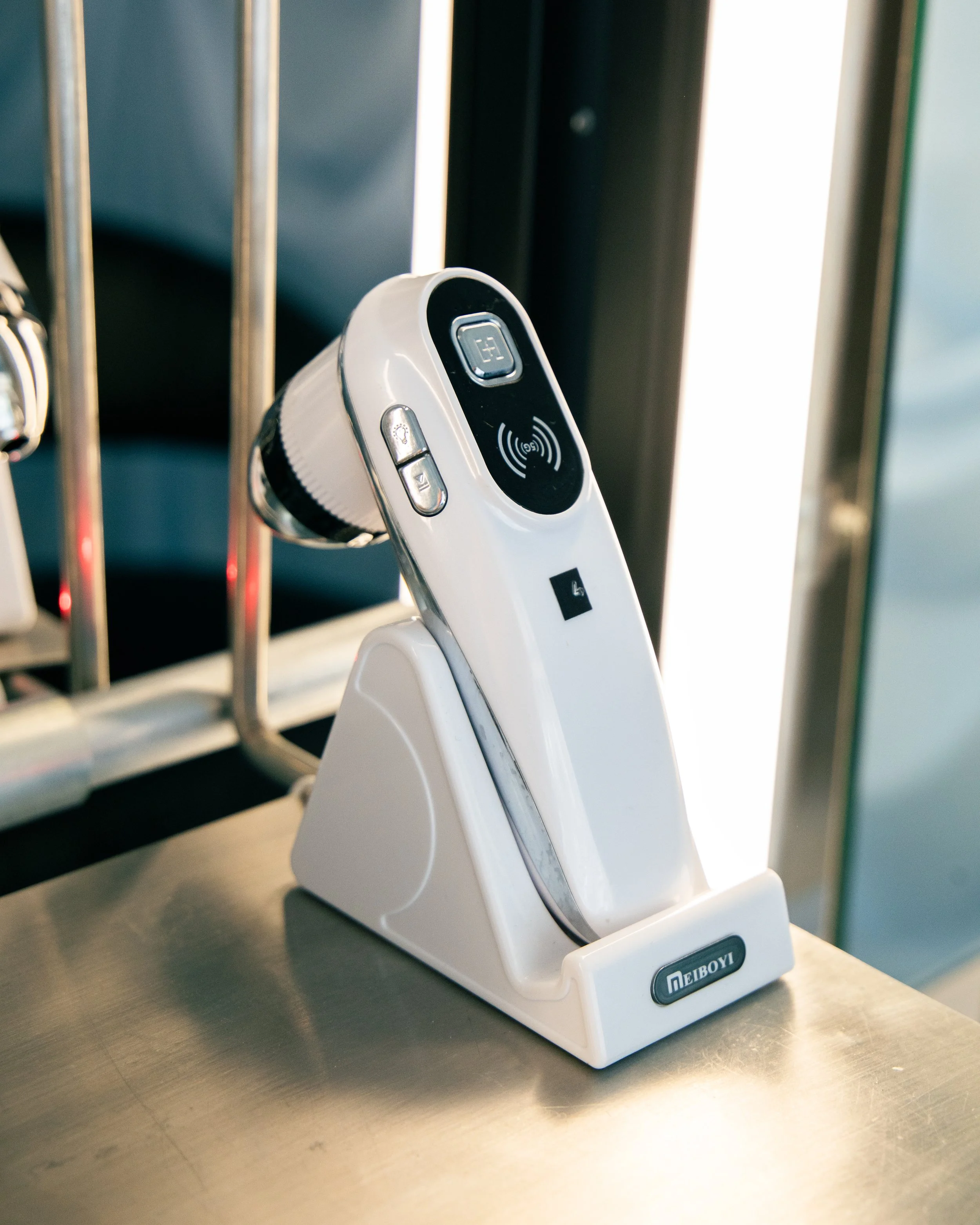 A white electronic key holder with a slot for a key fob, a circular button, and a wireless scan symbol, placed on a metallic surface in front of a metallic rack and vertical window.