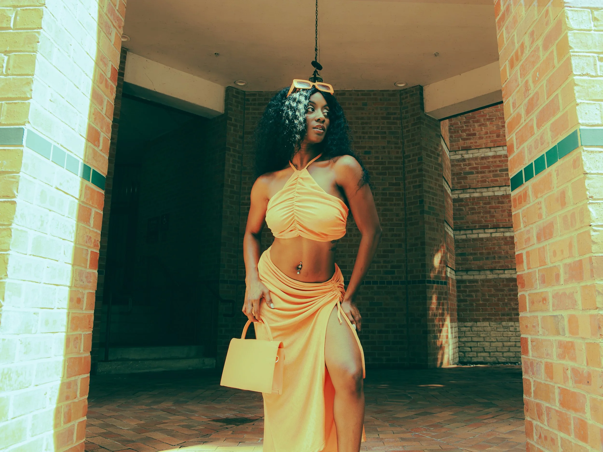 Local Dallas, Texas, DFW Non-traditional Fashion Editorial Creative Portraits Cover Art Photographer Photoshoot. A woman in a yellow dress standing outdoors at the entrance of a brick building, with sunglasses resting on her head and a yellow purse h