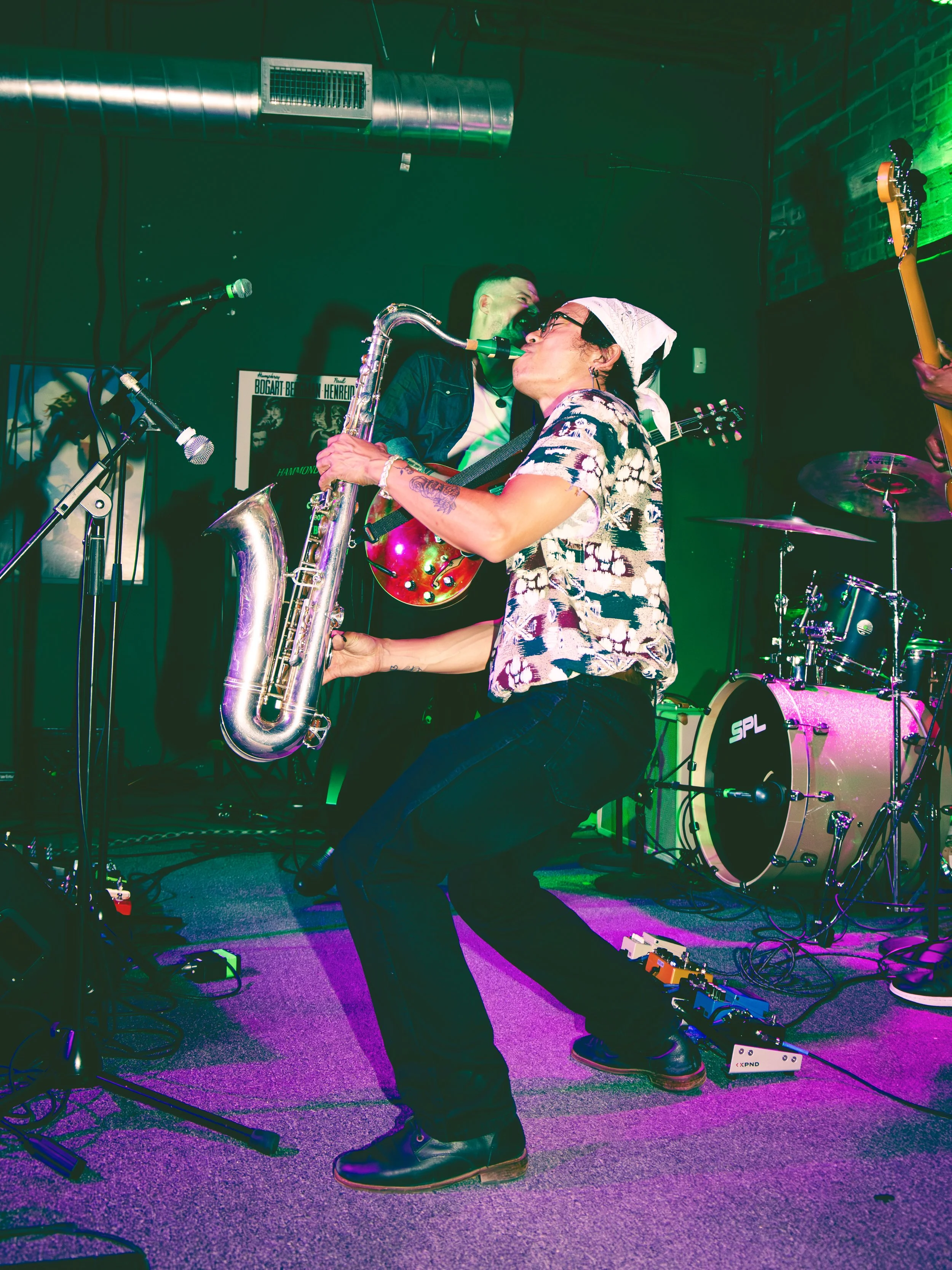 Musicians performing on stage with instruments including a saxophone, guitar, and drums under colorful lighting in a dark venue.