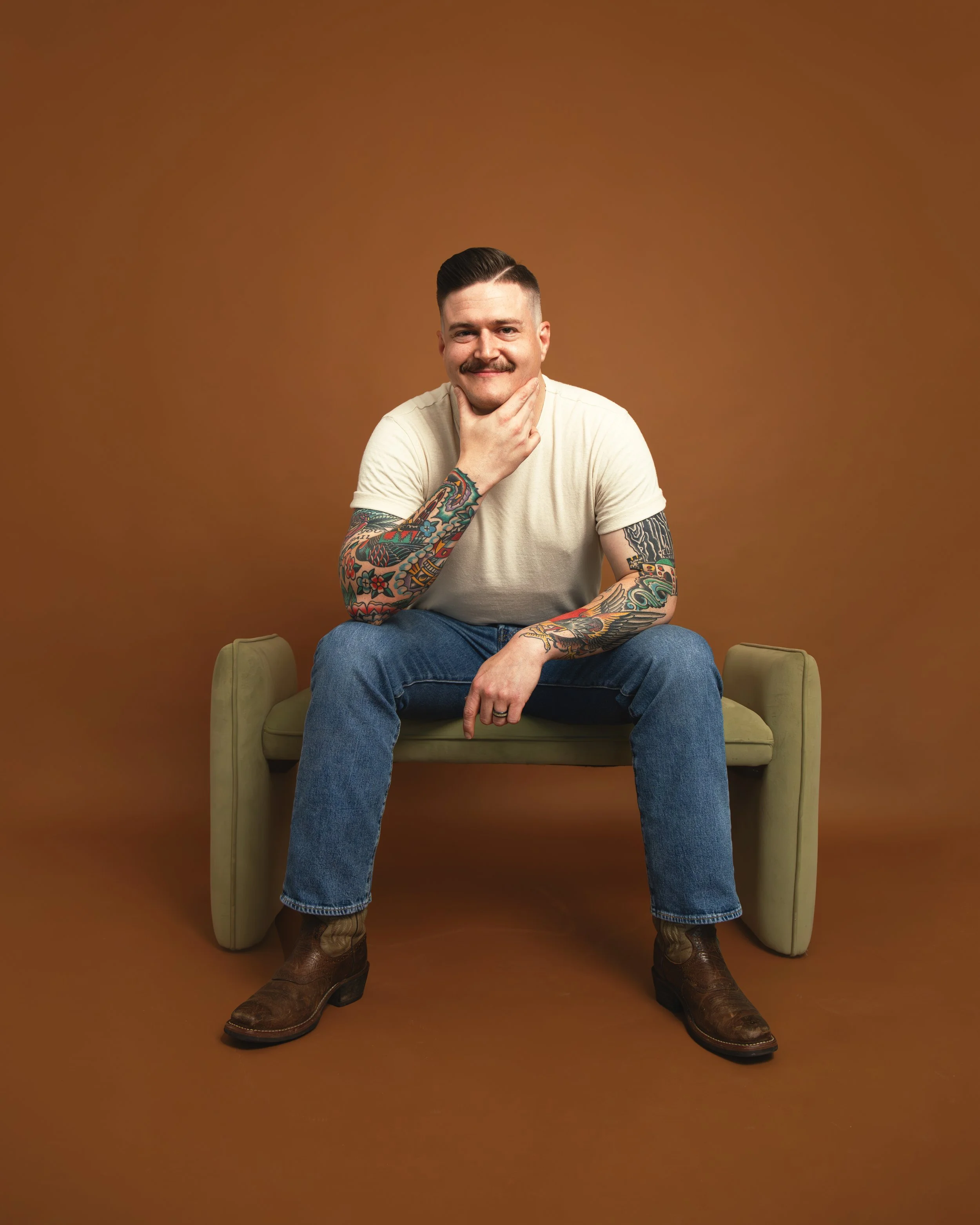 Local Dallas, Texas, DFW Non-traditional Fashion Editorial Creative Portraits Cover Art Photographer Photoshoot. Musician Caleb Rains Band . A man with tattoos on his arms, dressed in a white T-shirt and jeans, seated on a beige chair against a plain