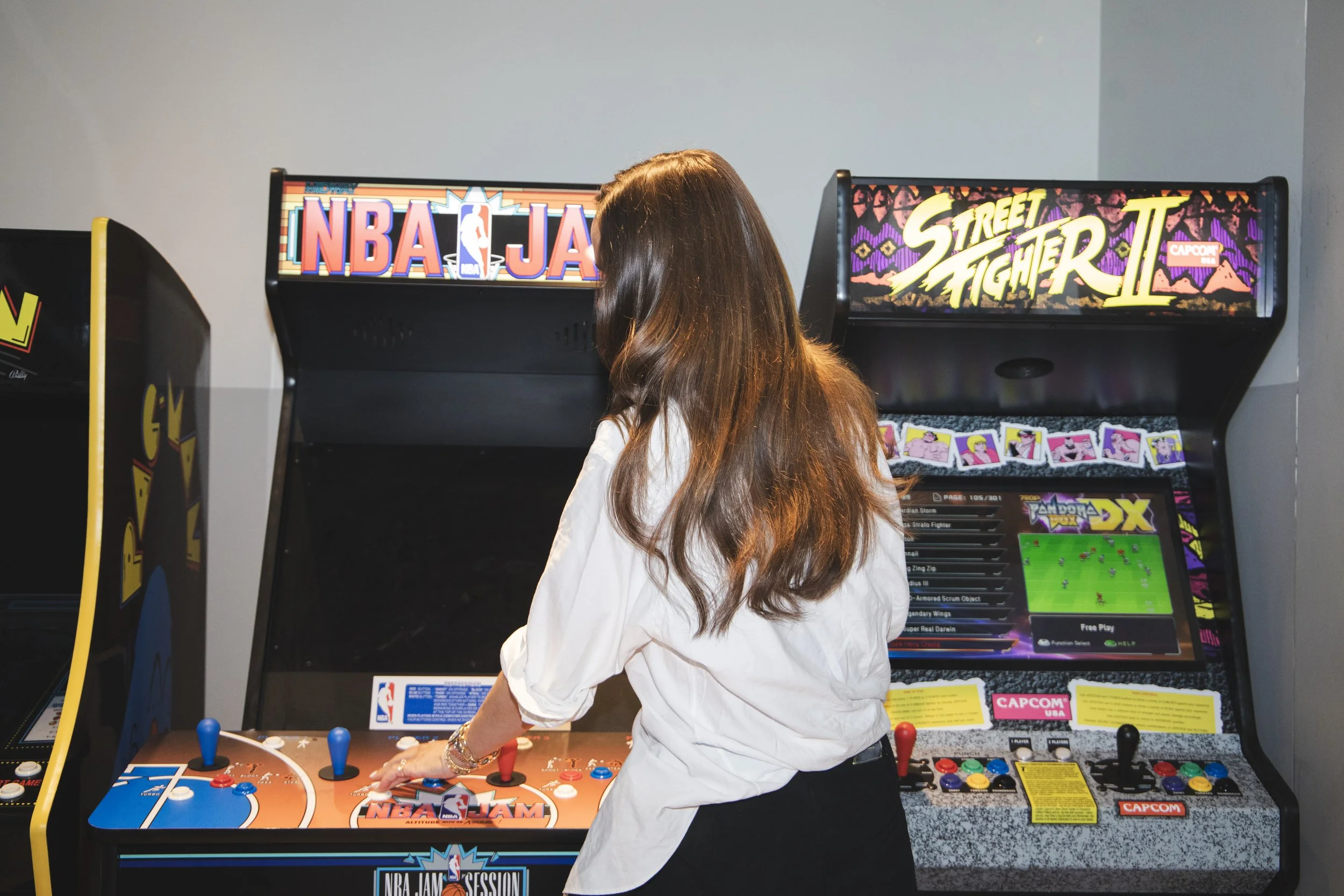 A woman with long brown hair wearing a white shirt is playing an arcade basketball game with a basketball hoop and backboard that is orange and blue.
