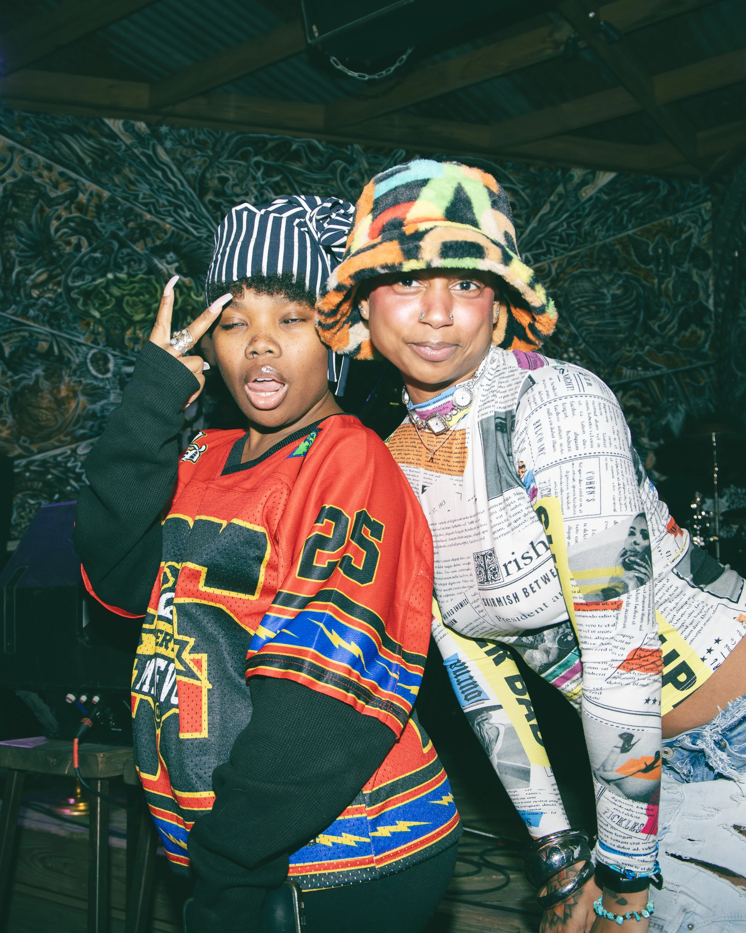 Two individuals posing in front of colorful and dark mural art, wearing vibrant, graphic clothing and accessories, with one person wearing a colorful bucket hat and the other a striped beanie, under a wooden ceiling with chain and chain link decorati