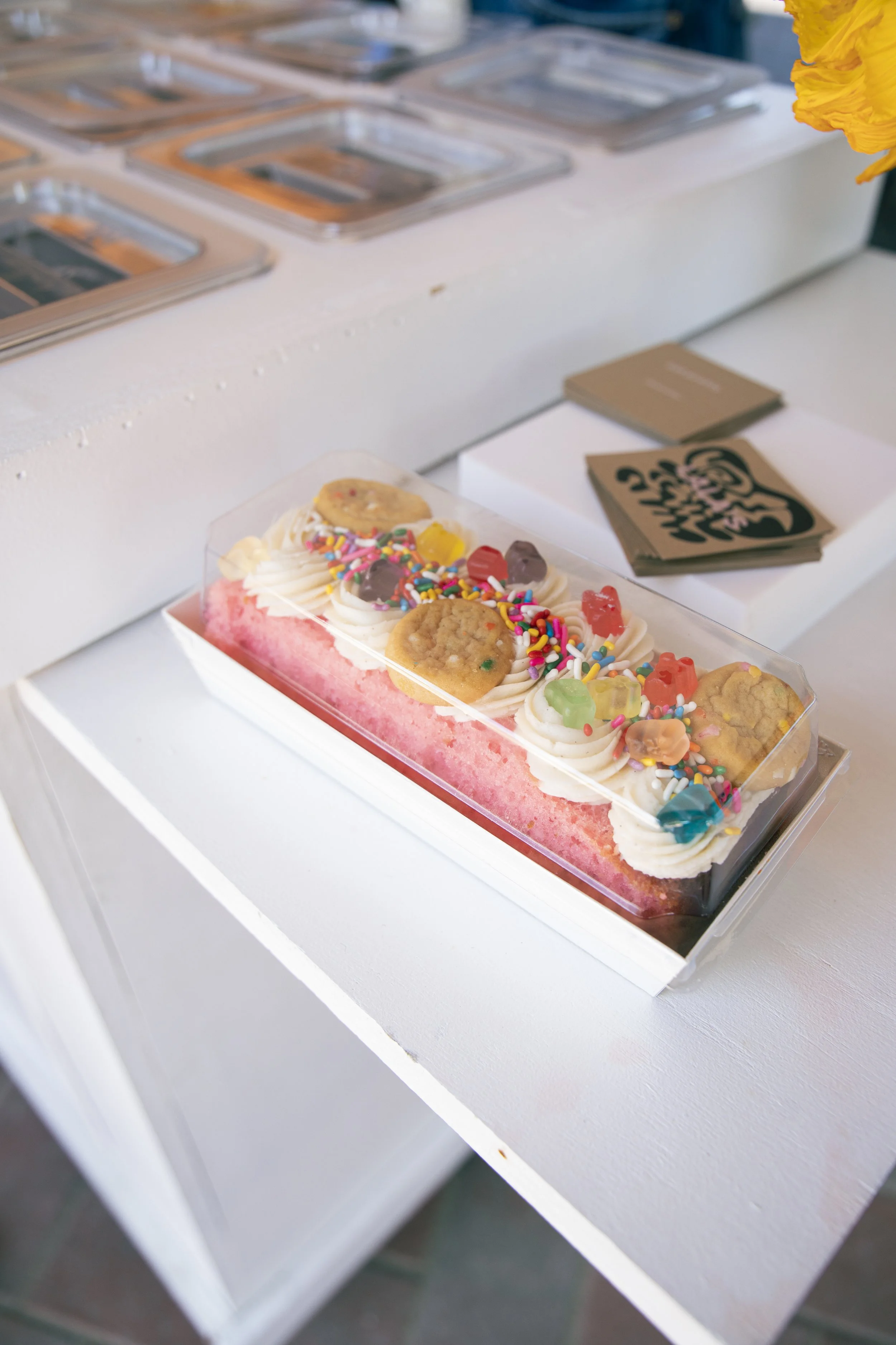 A colorful ice cream cake with sprinkles and cookies on top, placed in a clear plastic box on a white display surface.