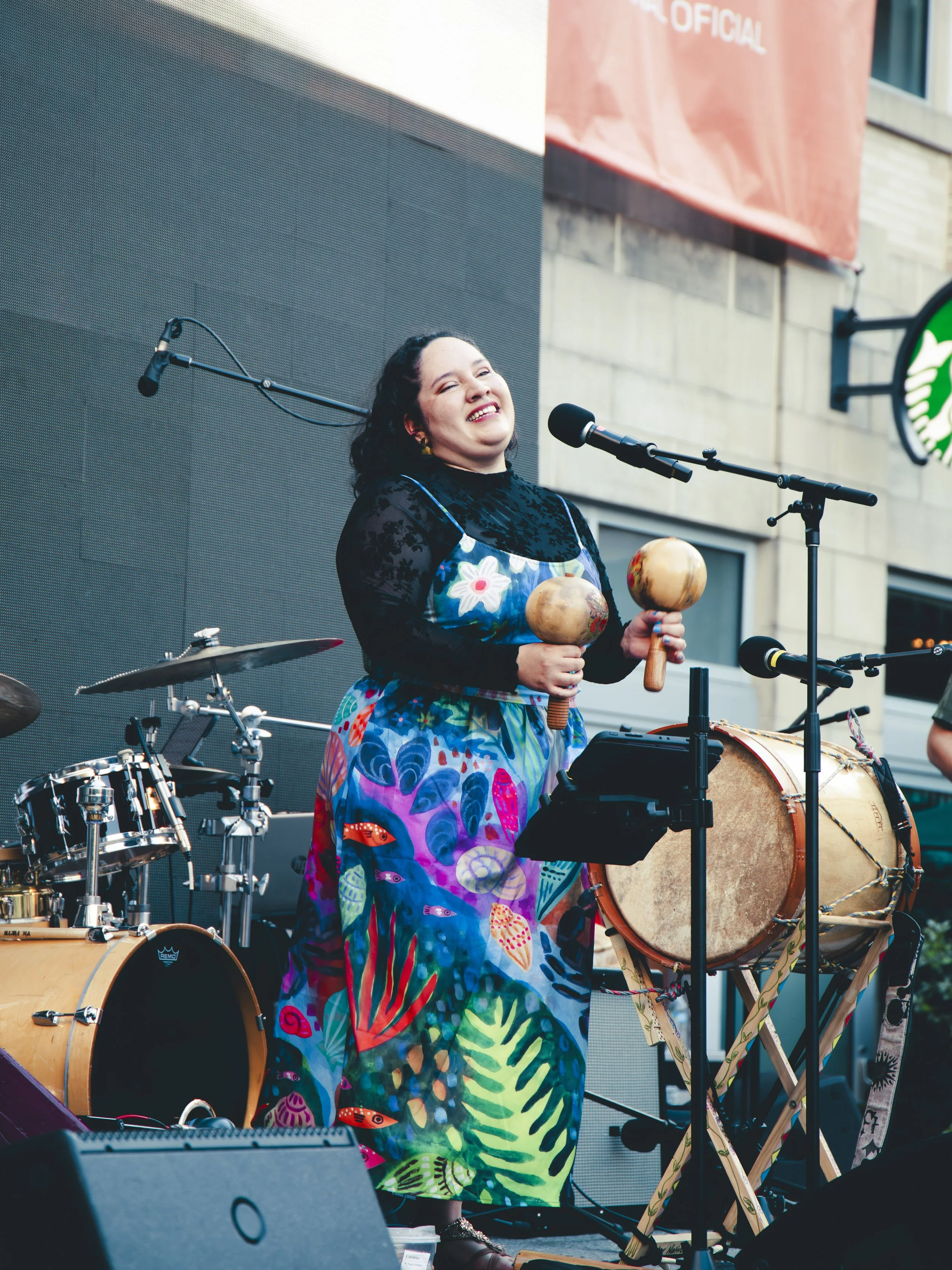 A woman wearing a colorful dress stands on stage, holding two maracas, smiling. She is surrounded by musical instruments including drums and drums, with microphones set up in front of her. There are background buildings and a large screen or projecti