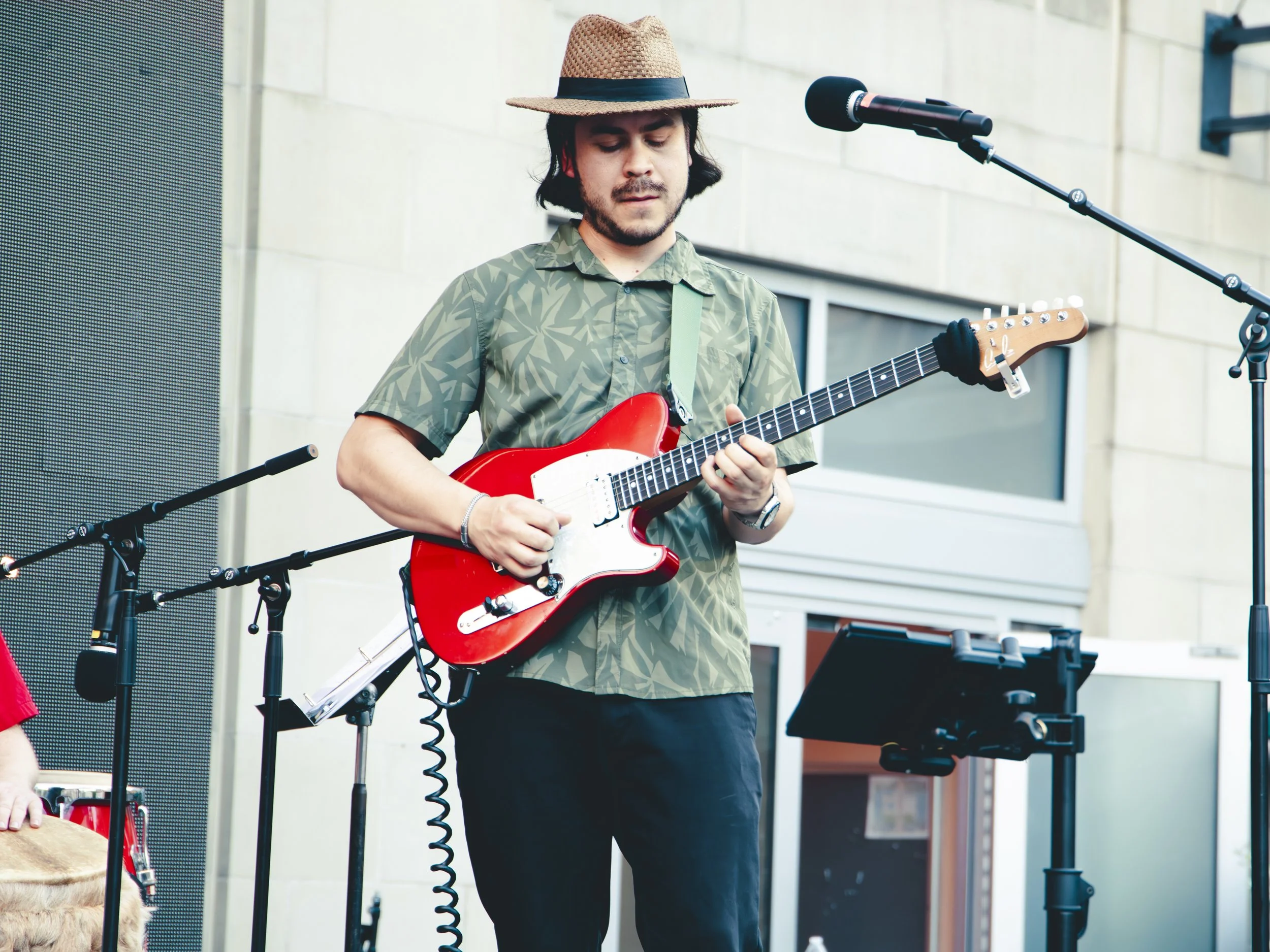 A man with dark hair and a beard playing an electric guitar on an outdoor stage, wearing a button-up shirt, black pants, a brown hat, and standing in front of microphones and speakers.